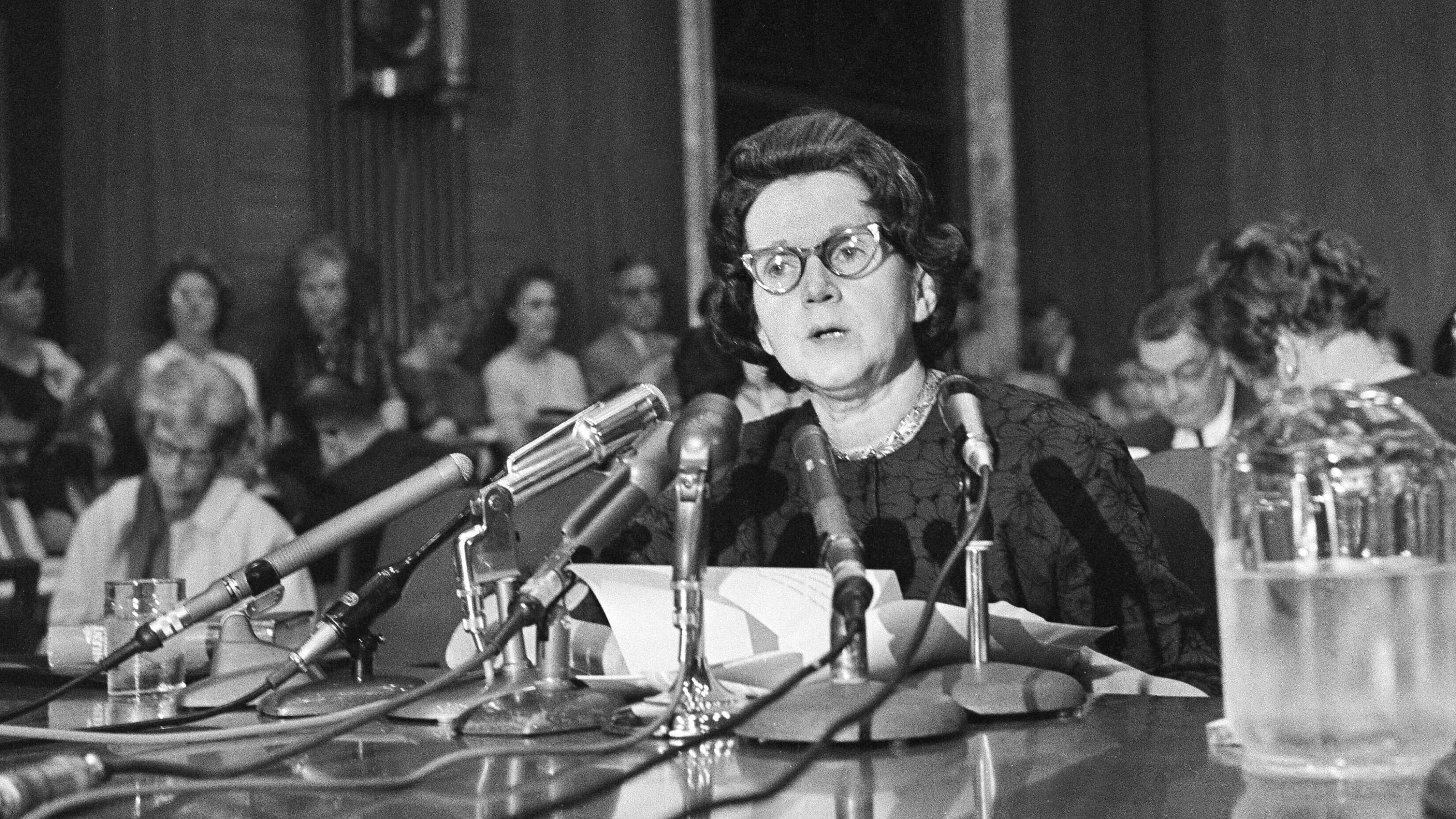 Schwarzweiss-Foto: Eine ältere Dame mit Brille sitzt in einem vollbesetzten Saal vor den Mikrophonen der Presse.
