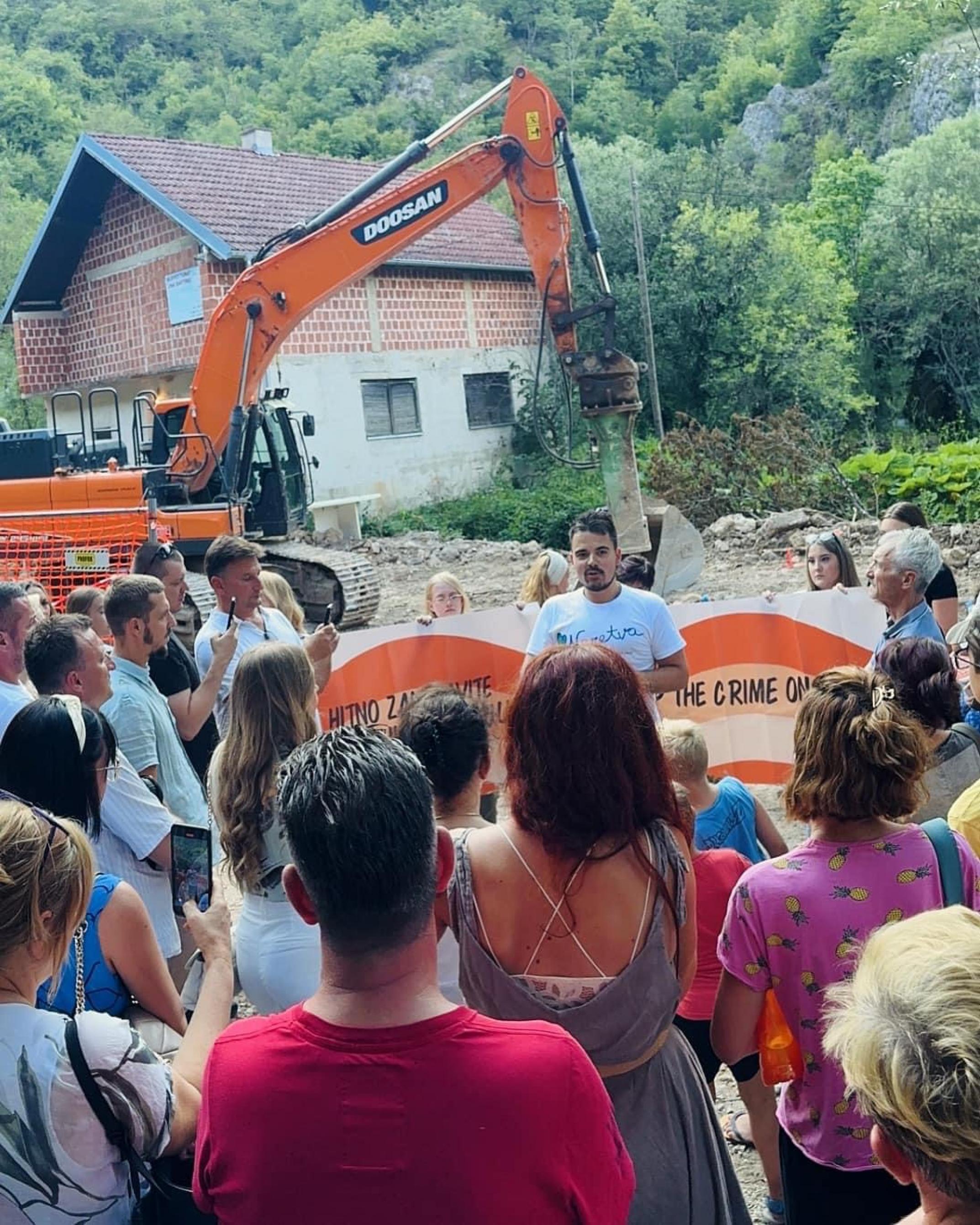 Mann in T-Shirt spricht vor Baustelle, Menschen hören im zu.