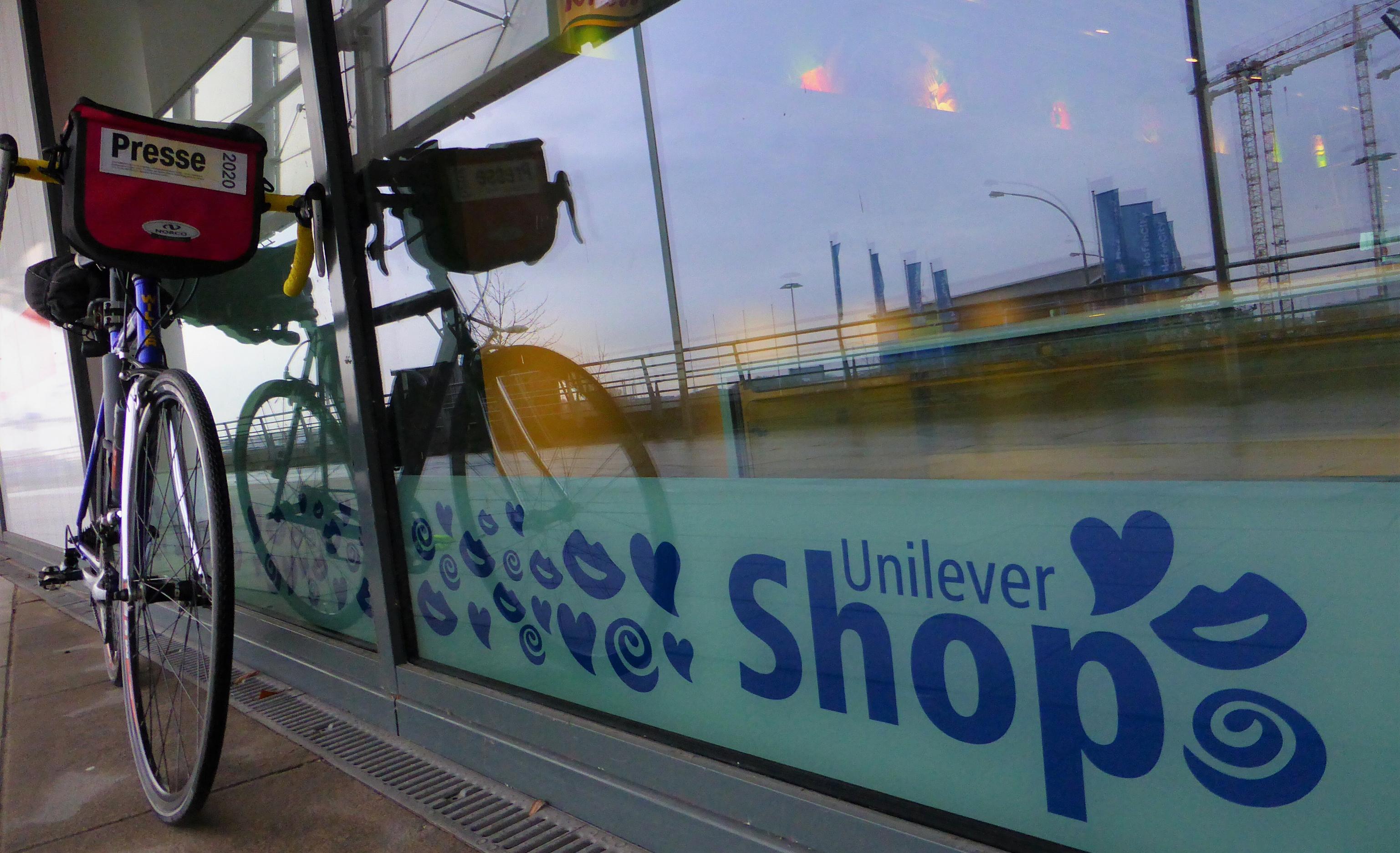 Unter einem Schaufensterglas, in dem sich an der Elbe befindliche Kraftwerkstürme und Baukräne spiegeln, wirbt eine mit Herzen und anderen Symbolen verzierte Werbeaufschrift für den sogenannten Unilever-Shop.