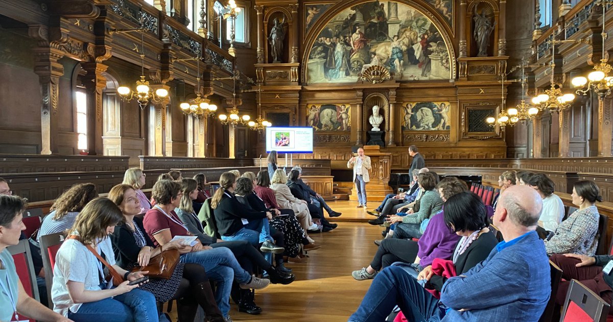 In einem holzgetäfelten Saal mit Büsten und Gemälden sitzen sich Menschen in drei Stuhlreihen gegenüber. Vorne redet ein Mann, neben ihm eine Beamerleinwand.
