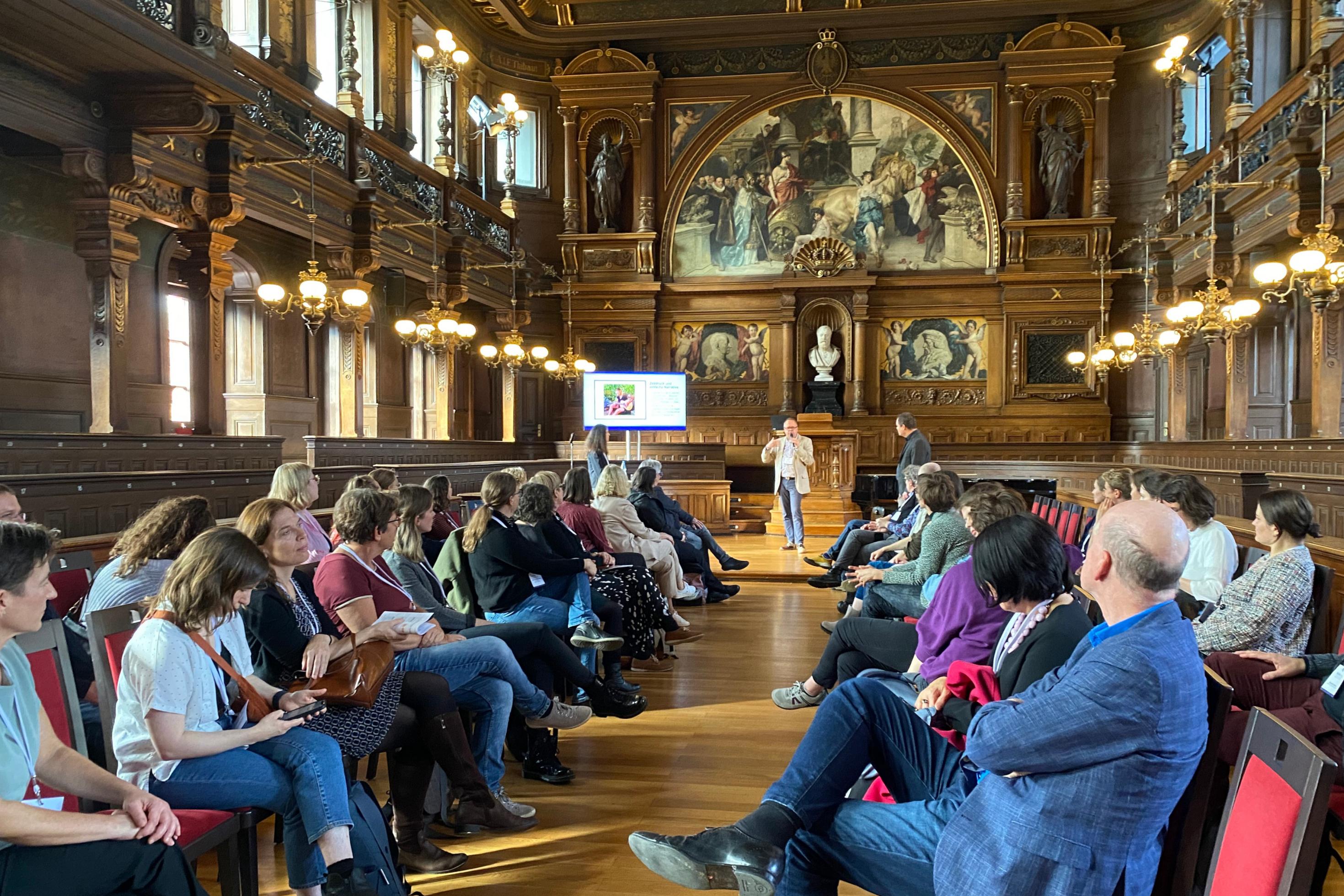 In einem holzgetäfelten Saal mit Büsten und Gemälden sitzen sich Menschen in drei Stuhlreihen gegenüber. Vorne redet ein Mann, neben ihm eine Beamerleinwand.