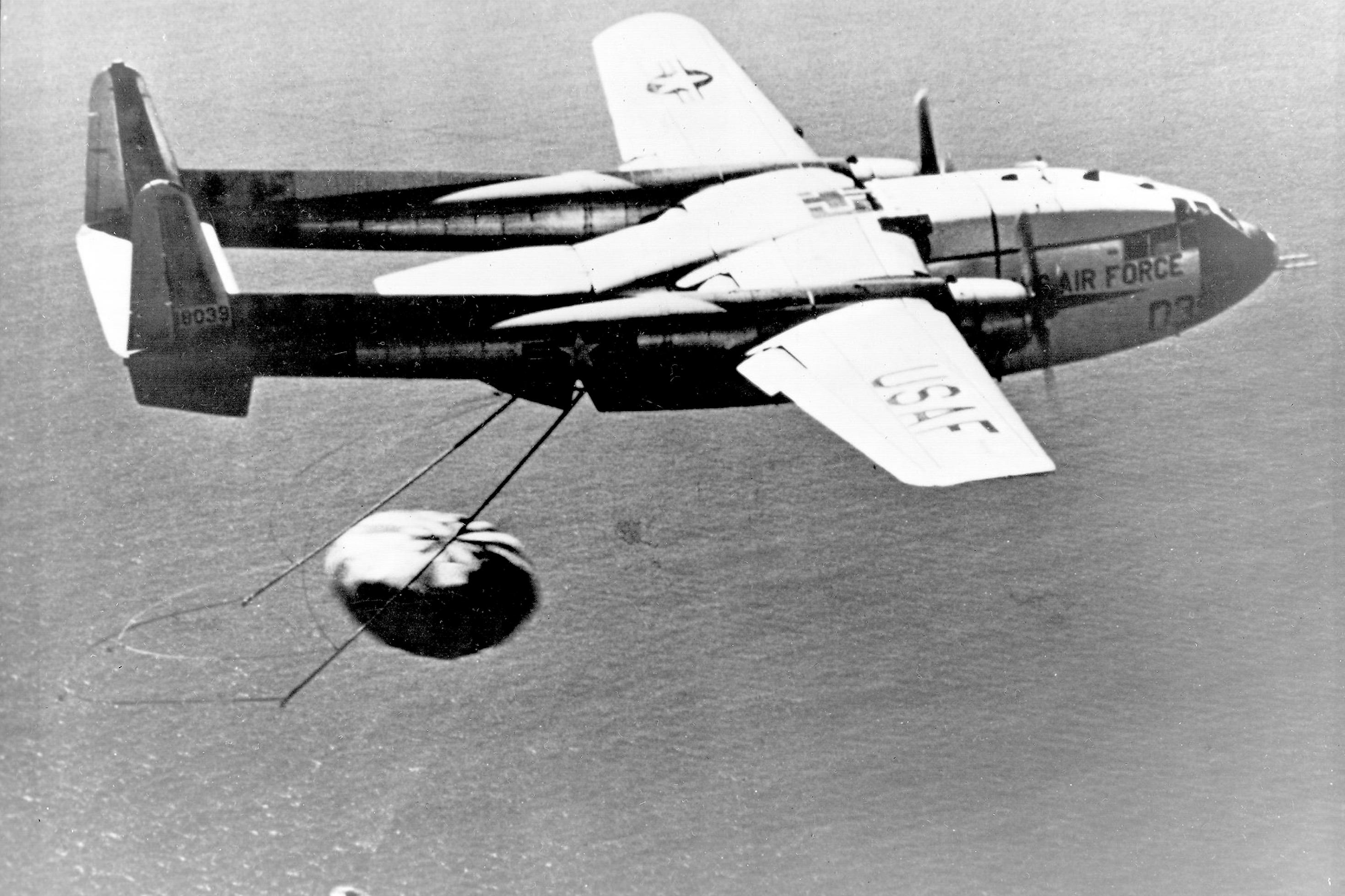 Ein fliegendes Propellerflugzeug über dem Meer, mit zwei über ein Seil verbundenen Stangen am Heck. Das Flugzeug fängt gerade eine Kapsel ein, die an einem Fallschirm schwebt.