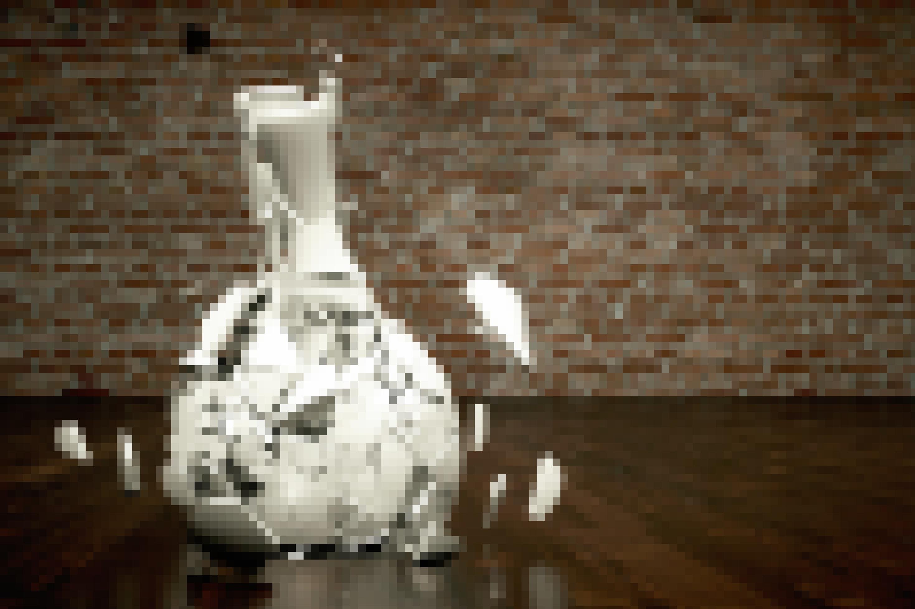 Zu sehen ist eine Vase aus weißer Keramik, die auf einen dunklen Holzfußboden prallt und dabei in zig Bruchstücke zerbirst. Im Hintergrund eine Wand aus rötlichen Mauersteinen.