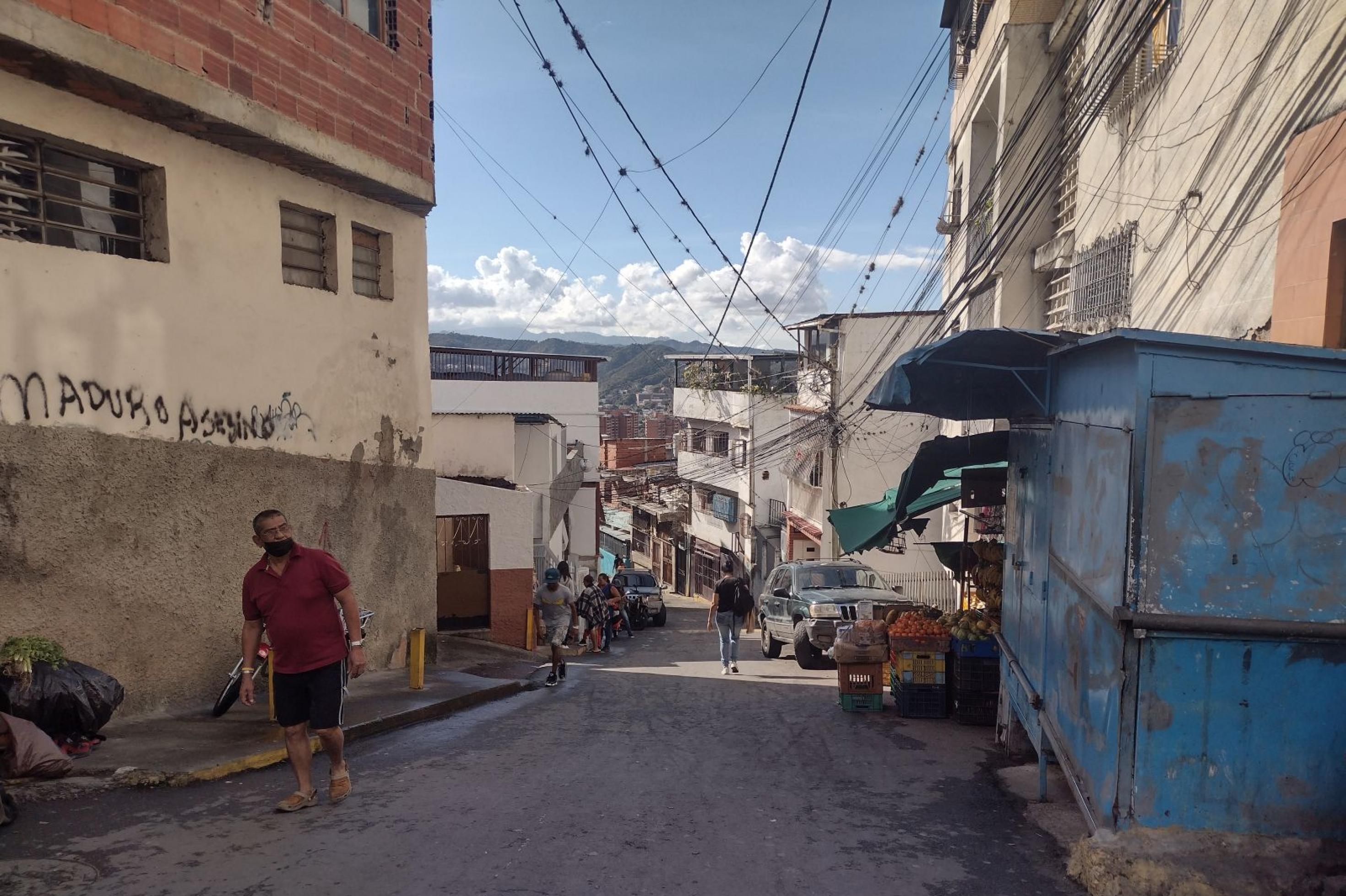 Enge Straße mit Blick auf Caracas, rechts und links ärmliche Häuser, darüber Stromleitungen. Ein Mann in kurzen Hosen läuft die Strasse hoch. An der Wand ein Graffiti: Maduro Mörder.