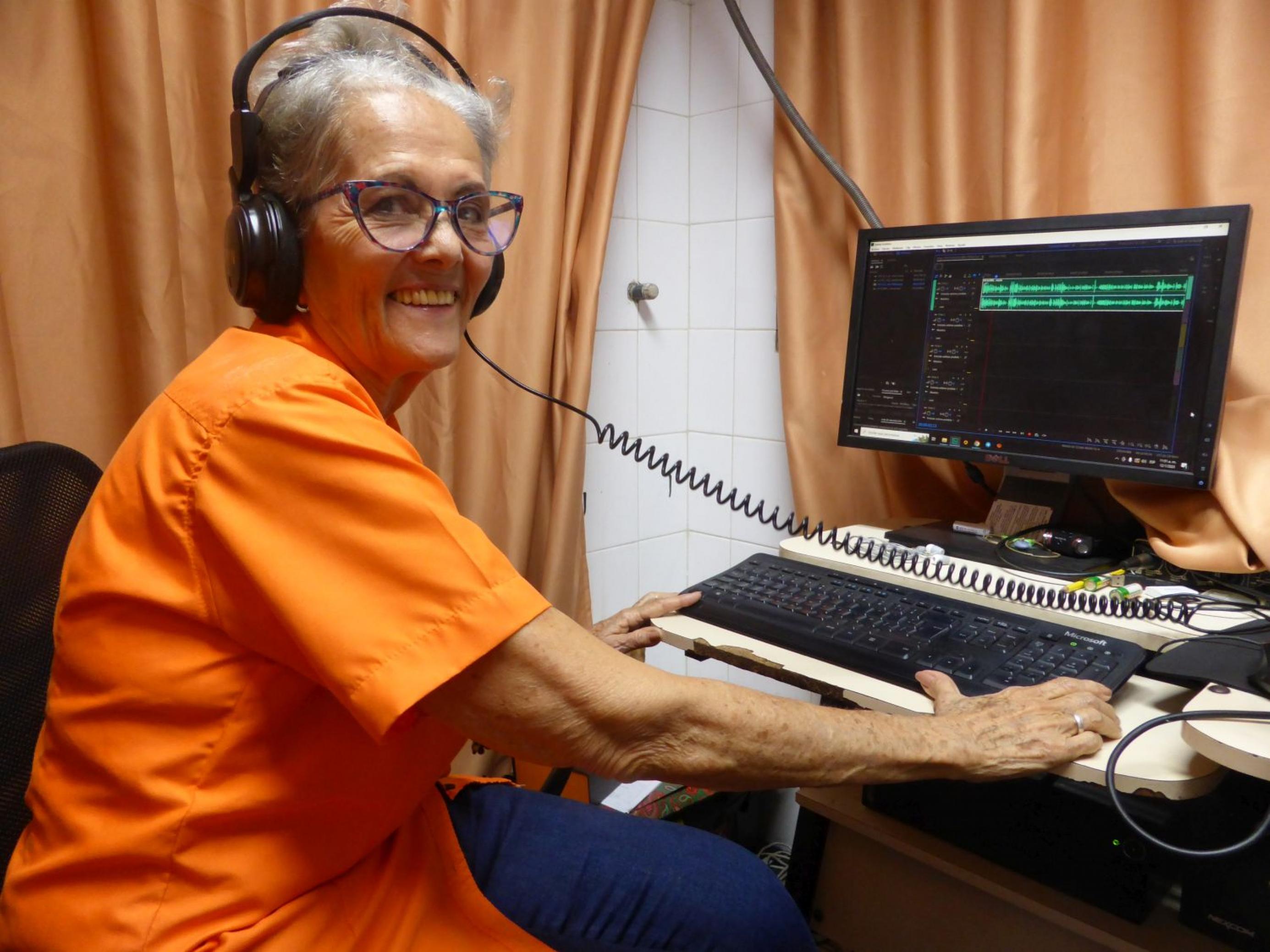 70-jährige Frau mit kurzen grauen Haaren, orangener Bluse, Kopfhörer vor einem PC sitzend, lacht in die Kamera.