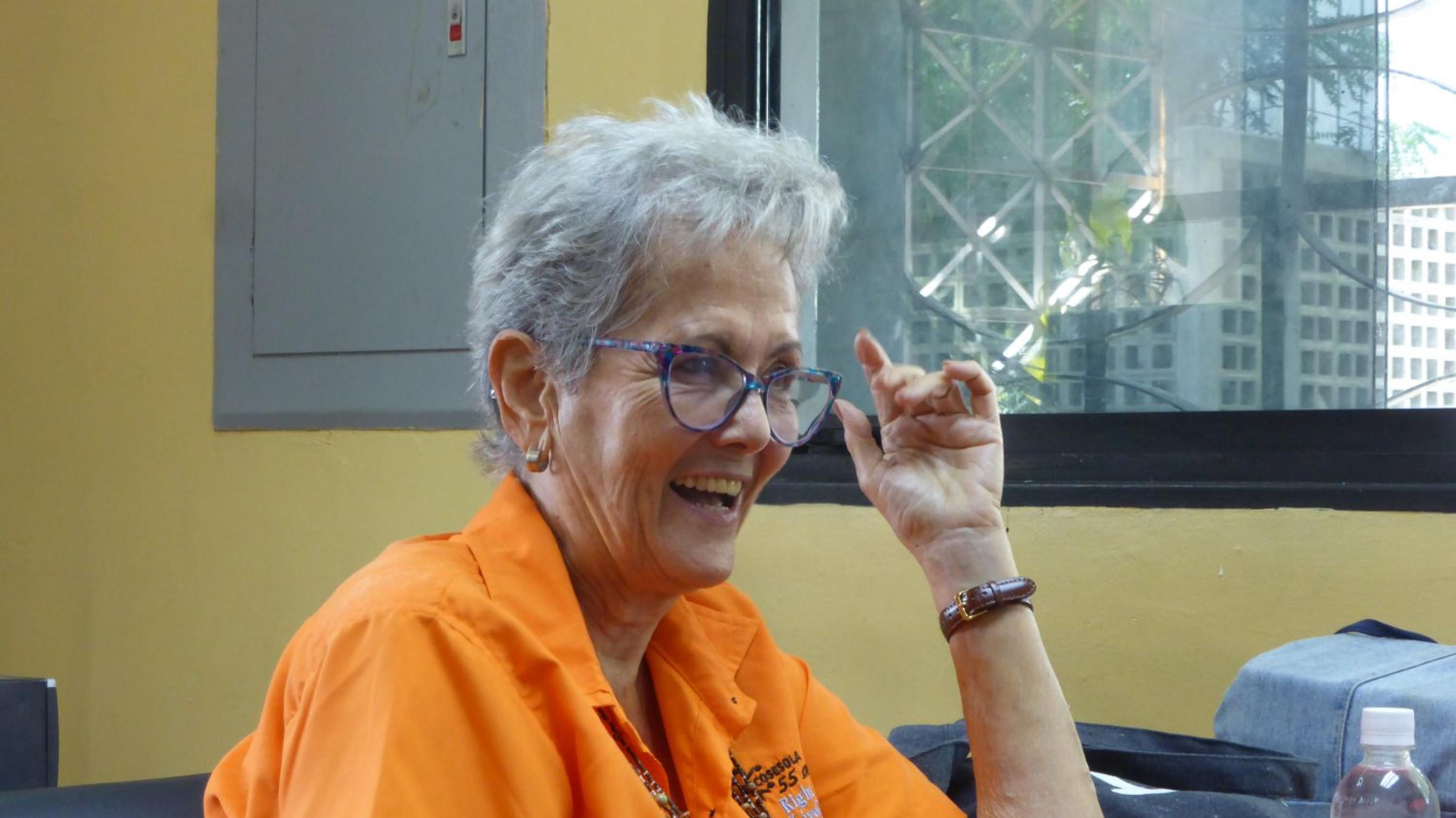 Frau, 70 Jahre alt, kurzes graues Haar, Brille, kurzärmliges orangene Bluse, sitzt an einem Tisch und spricht.