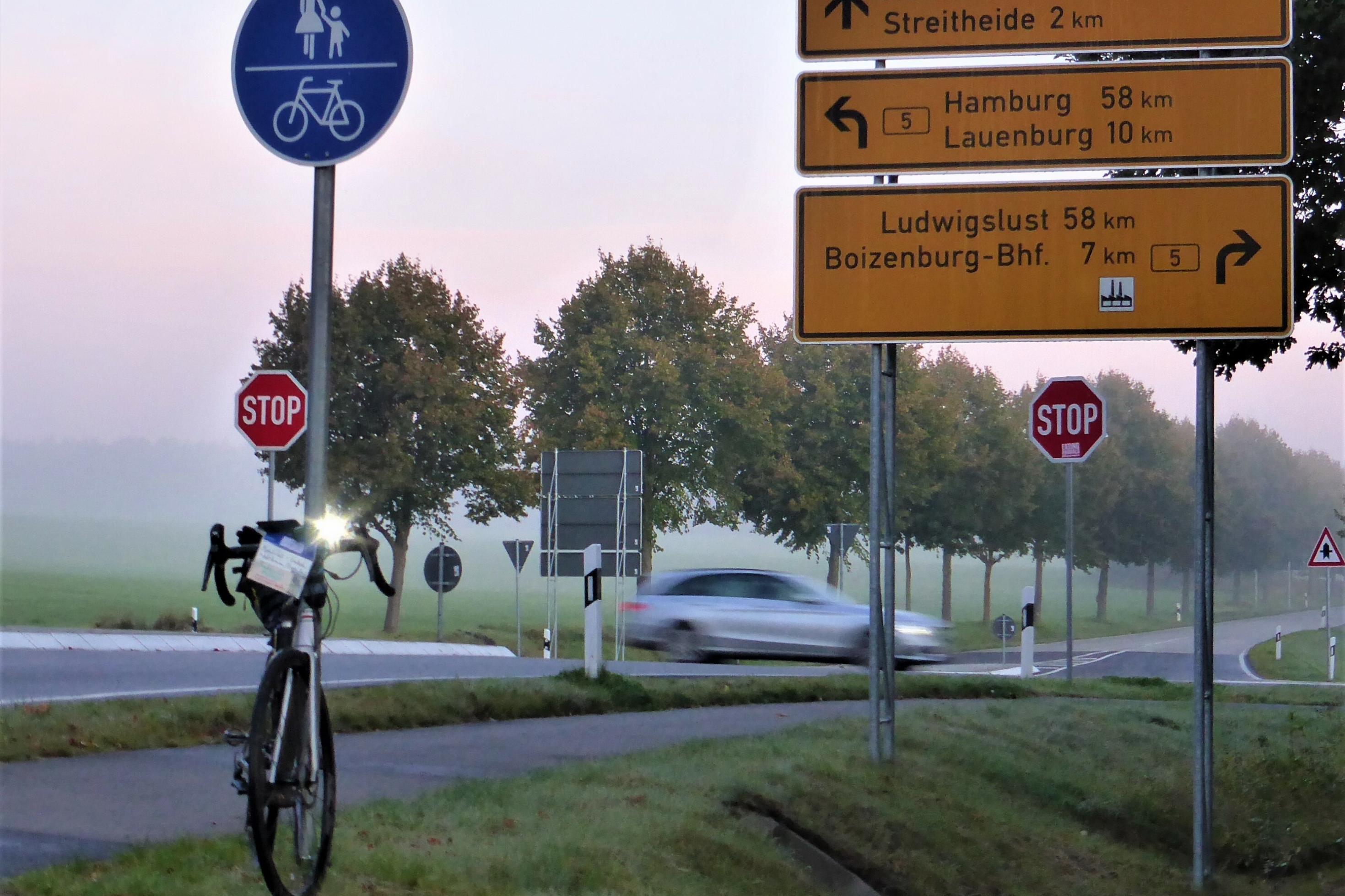 Das Rennrad des RadelndenReporters lehnt im fahlen Morgenlicht an einem Radwege-Schild, hinter dem Stoppschilder sowie ein vorbeirauschender Pkw zu sehen sind.