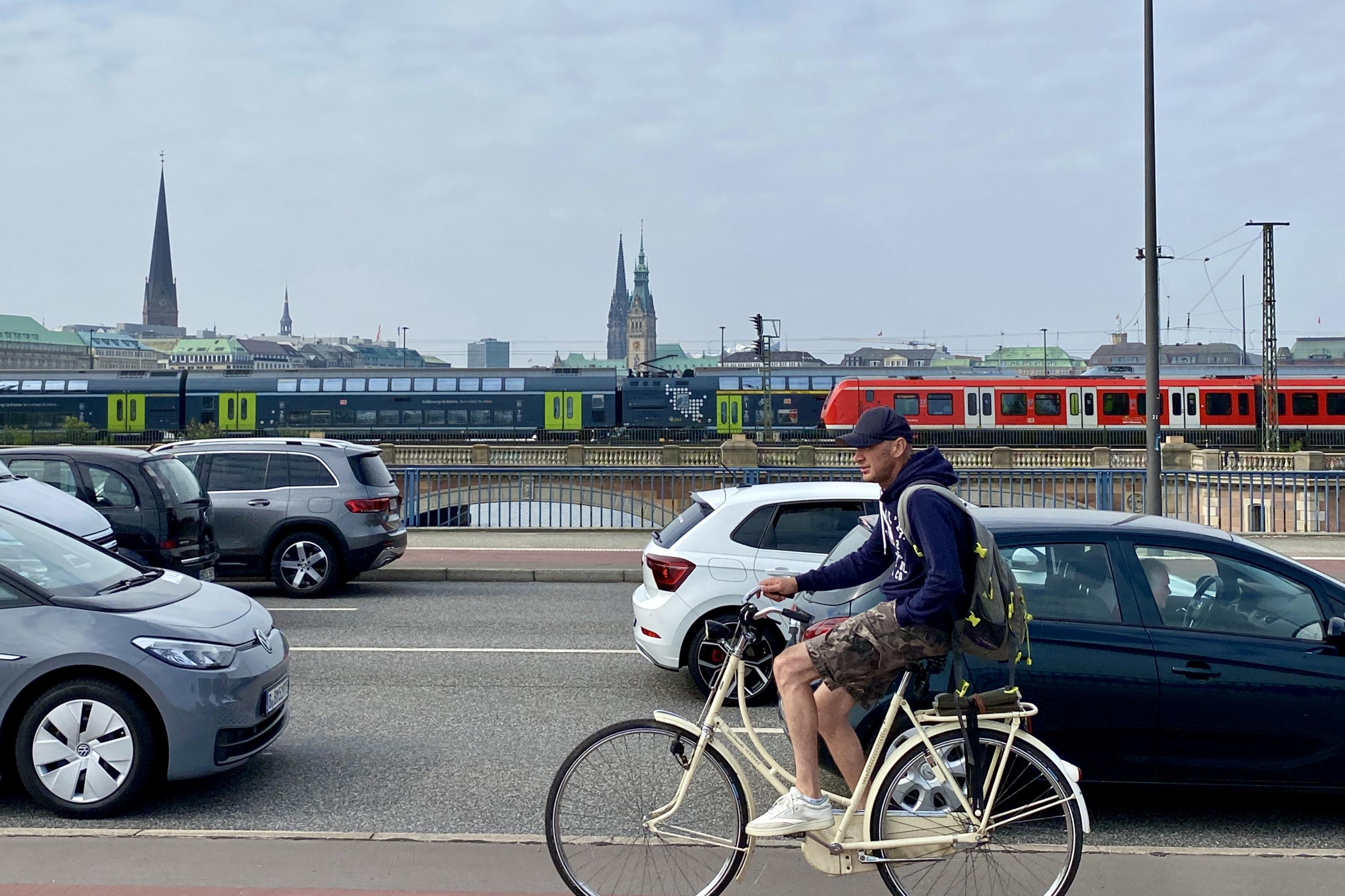 Ein Radfahrer fährt an Autos vorbei. Im Hintergrund fahren Bahn und S-Bahn.