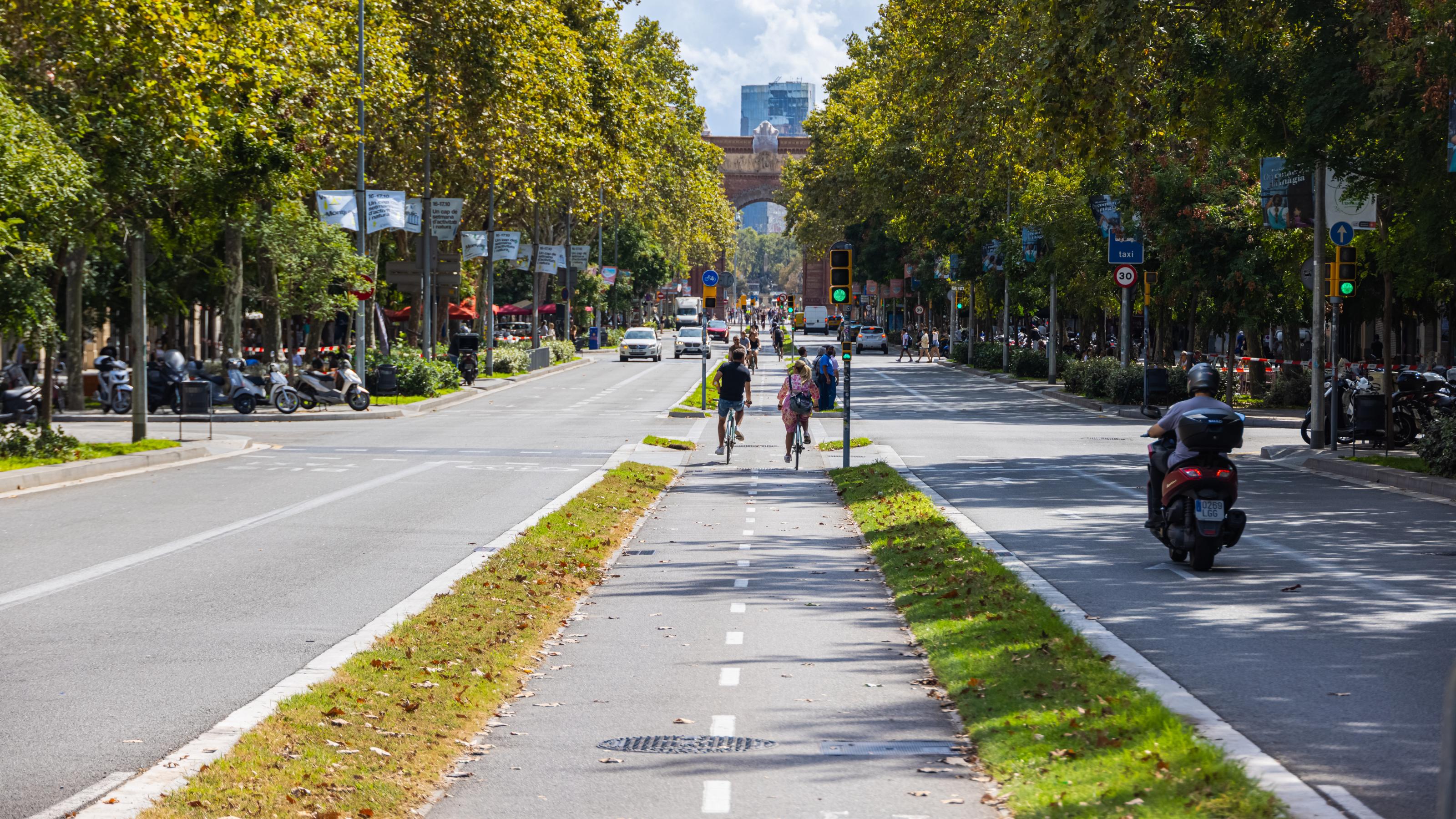 Inmitten einer von Bäumen gesäumten vierspurigen Straße sind zwei Radfahrende auf einem breiten Radweg unterwegs, der zusätzlich mit Grünstreifen von den Fahrbahnen links und rechts abgetrennt ist.