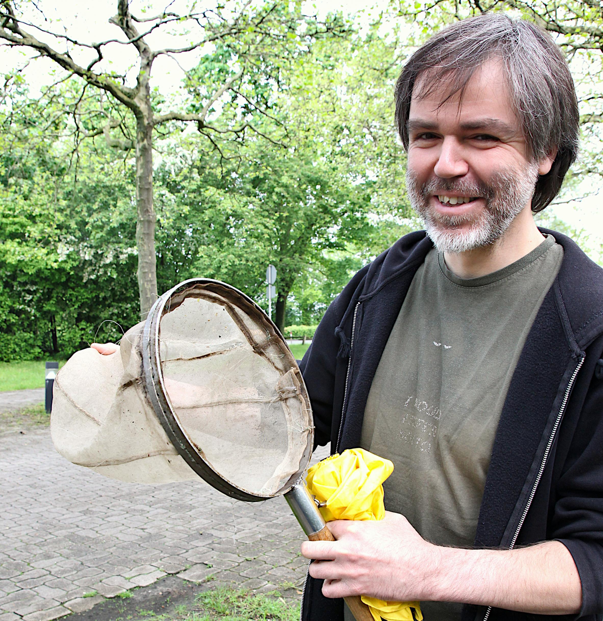 Ein Mann mit oliv-grauem T-Short und dunkelblauer Hoodie-Jacke lächelt in die Kamera. Er hält einen Kescher sowie einen zusammengefalteten, gelben Regenschirm in der Hand.