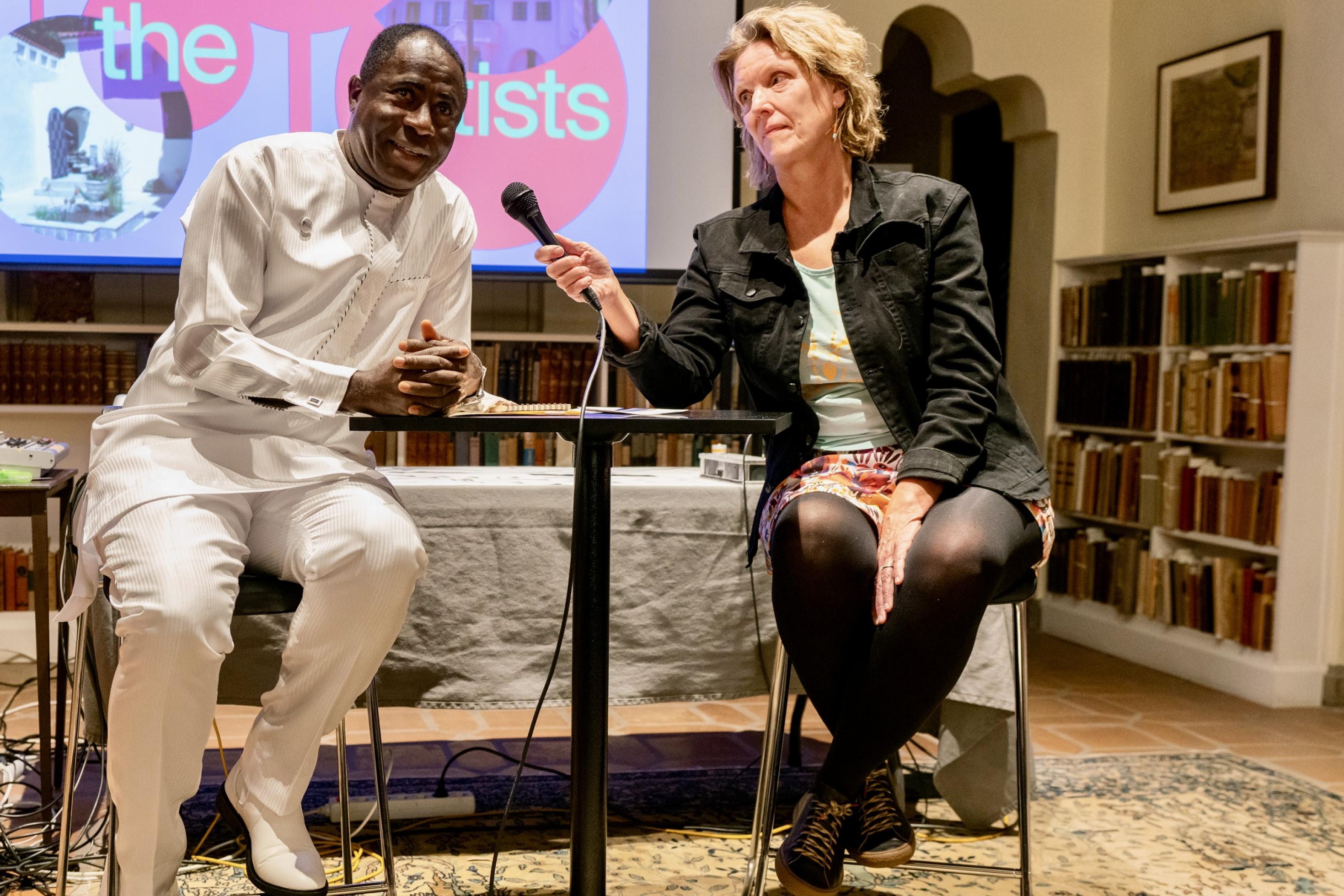 Der nigerianische Journalist Eric Ojo im Gespräch mit einer Journalistin. Sie sitzen sich an einem Tisch gegenüber, dem Publikum zugewandt, im Hintergrund eine Projektion mit der Aufschrift „Meet The Artists“
