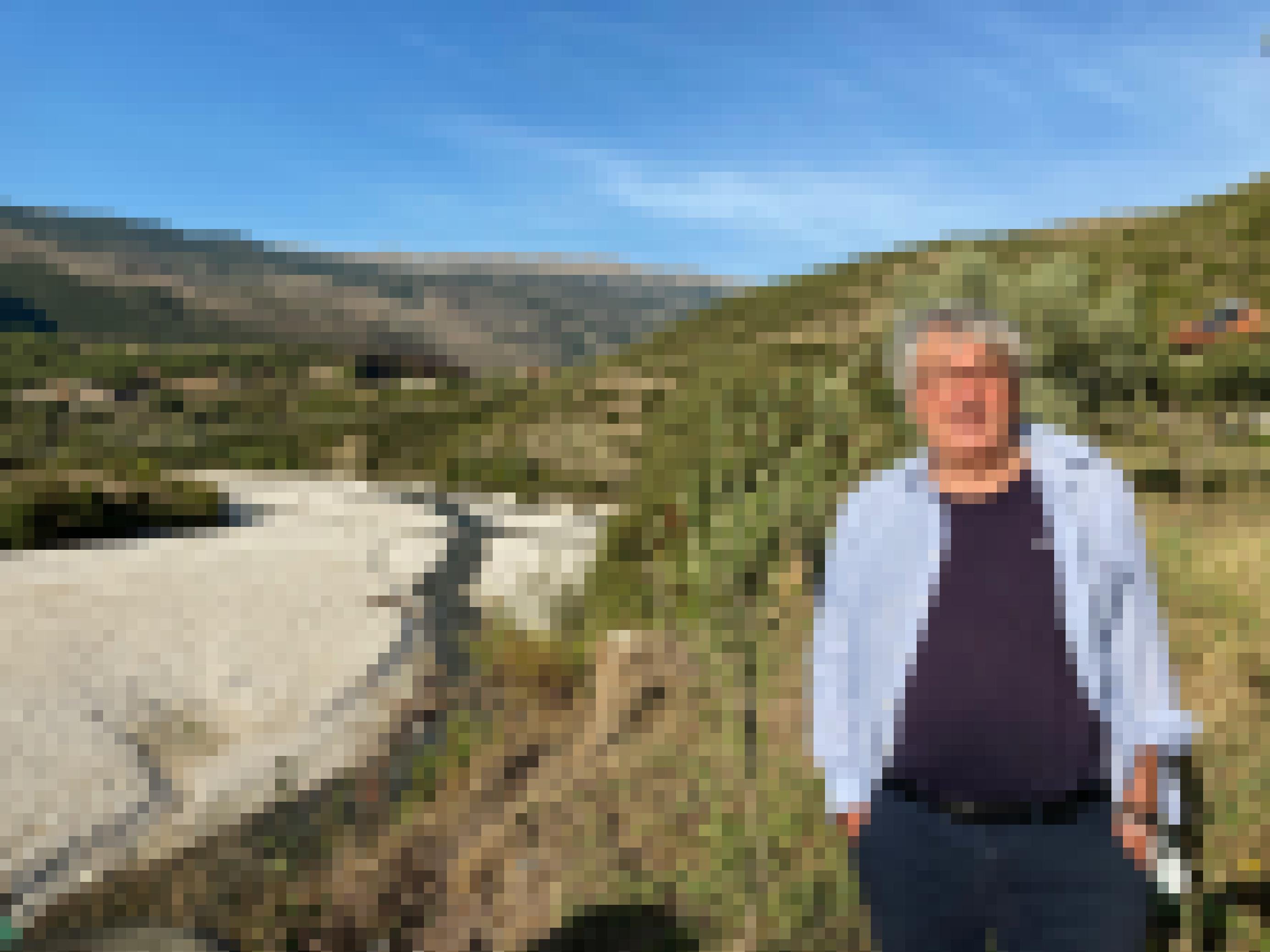 Mann mit grauen Haaren in einem T-Shirt und Hemd steht auf einem Hügel, im Hintergrund sieht man den breiten Fluss und Berge.