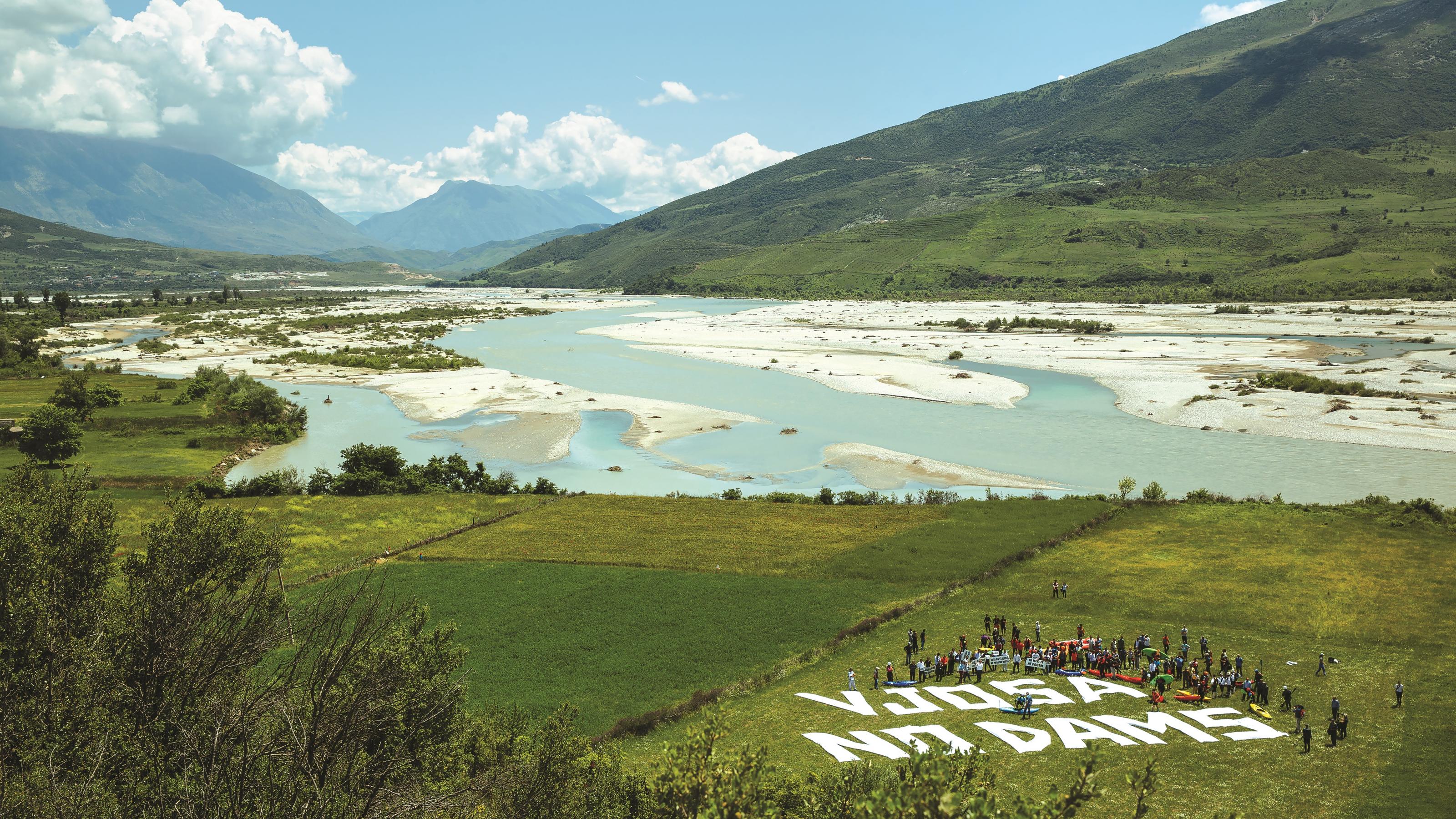 Flusslandschaft mit türkisfarbenem Wasser, große Schotterbänke, weiße Buchstaben auf der Wiese: Vjosa no dams