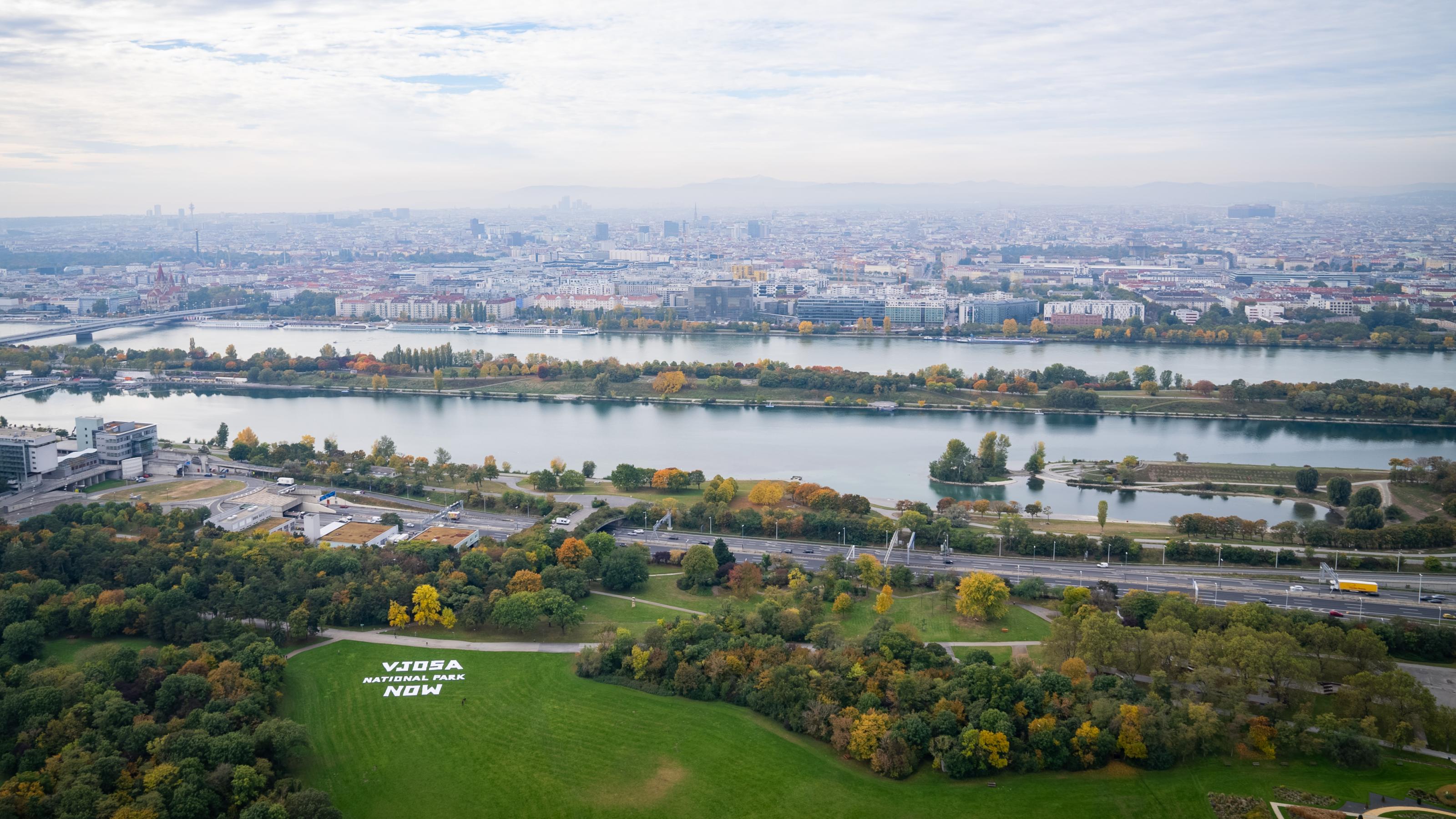 Der Fluss Donau mit der Stadt Wien von hoch oben fotografiert, unten auf der Wiese ein weißer Schriftzug.