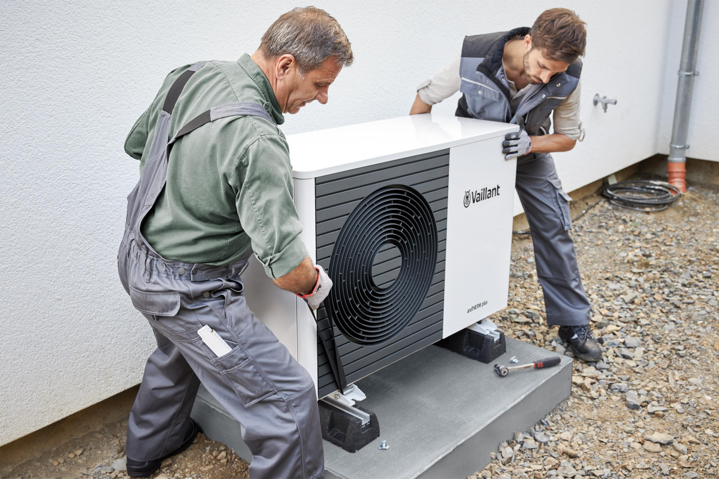 Zwei Handwerker in Arbeitshosen stellen eine Wärmepumpe auf eine Betonplatte neben einer Hausfassade.