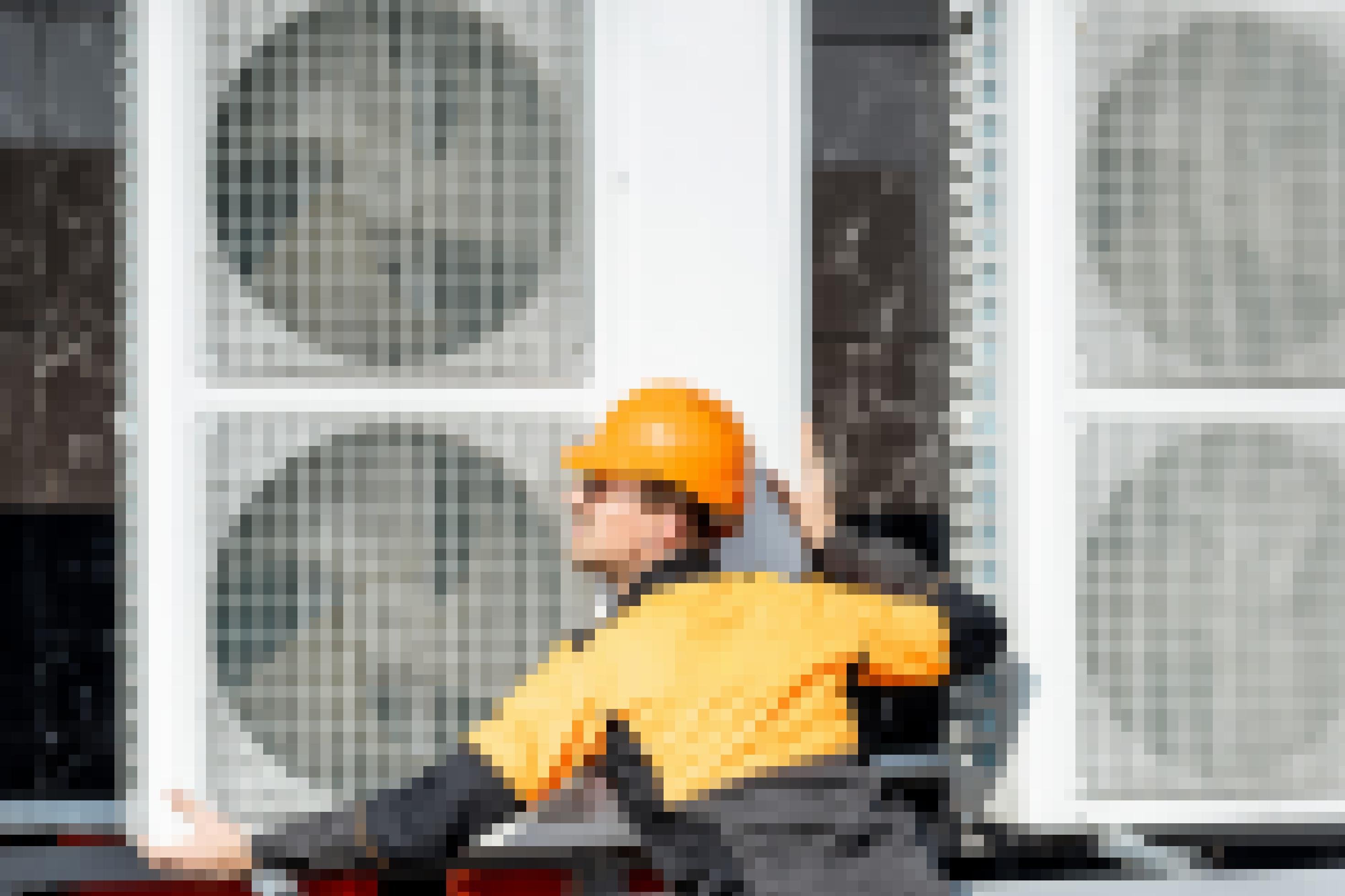 Ein Mann mit orangener Jacke und Bauhelm installiert einen Kasten mit Ventilatoren an einer Hauswand.