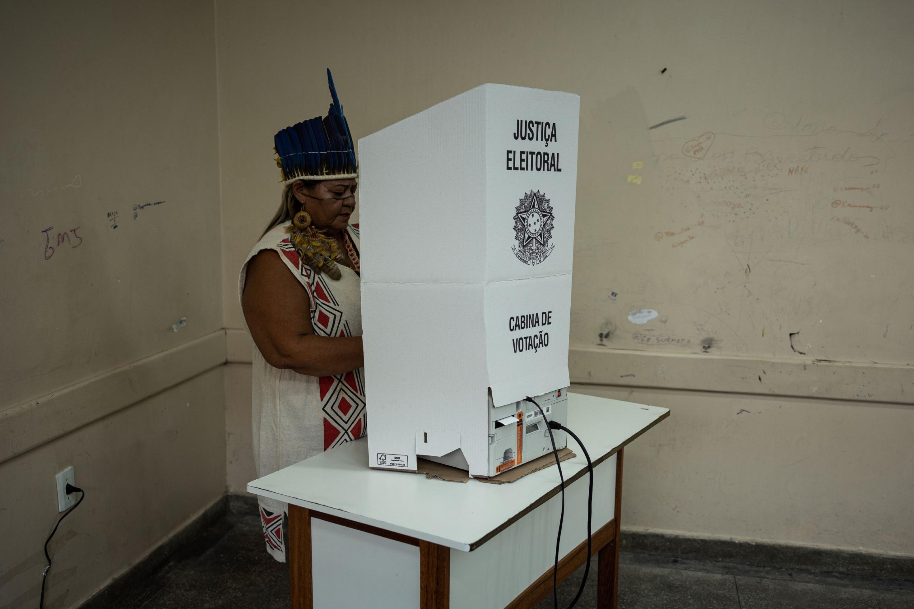 Eine mit Federschmuck bekleidete Frau steht hinter einem Schreibpult, auf dem ein Pappe-Aufsatz über einem elektronischen Wahlgerät das Wahlgeheimnis schützen soll.