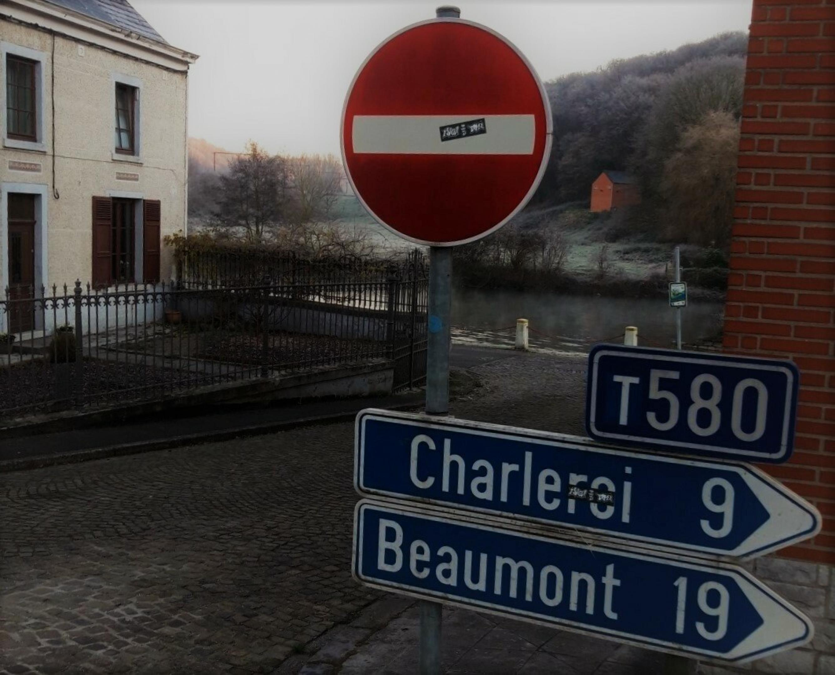 Ein blaues Straßenschild gibt die Entfernung nach Charleroi – in weißer Schrift – mit 9 Kilometern an. Hinter dem Schild stehen Häuser, fließt ein Fluss und breitet sich rau bereifter Wald, auf den eben die ersten Sonnenstrahlen fallen.