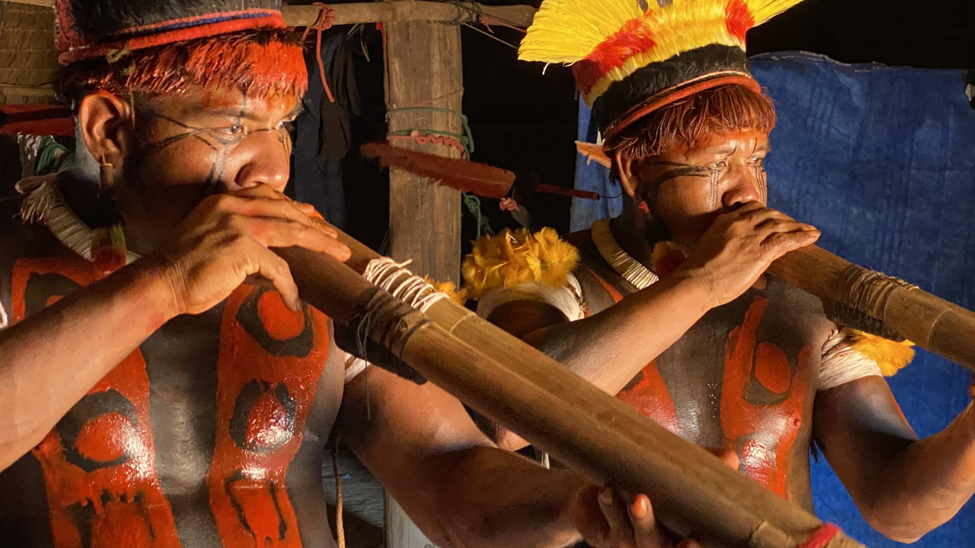 Zwei mit roter Farbe bemalte und mit Federn geschmückte Mehinako-Indigene spielen ihre Flöten im Duett. Sie tanzen von Haus zu Haus und rufen das Dorf in einen mythischen Zustand.