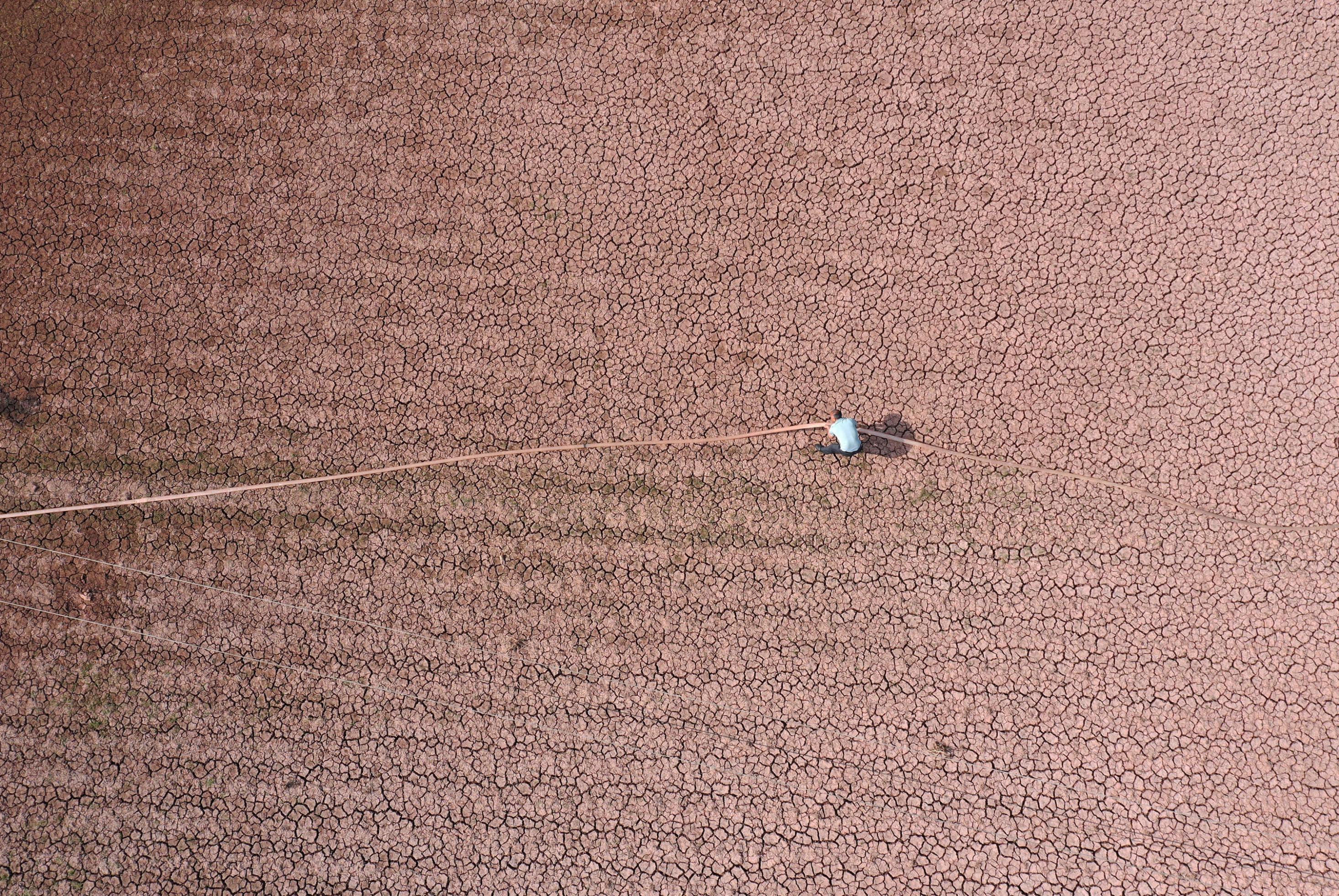 Luftaufnahme eines ausgetrockneten Feldes mit rissigem Boden, auf das ein Bauer einen Wasserschlauch zieht.