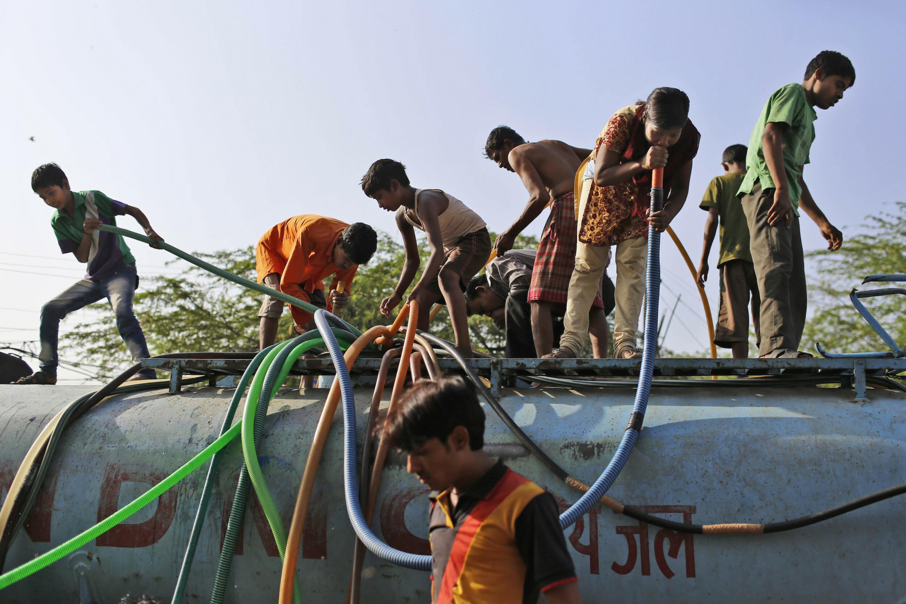 Jugendliche stehen auf einem Tanklastwagen mit Wasser und reichen Schläuche nach unten zum Befüllen von Behältern. Ein Mädchen trinkt Wasser direkt aus einem der großen Schläuche.