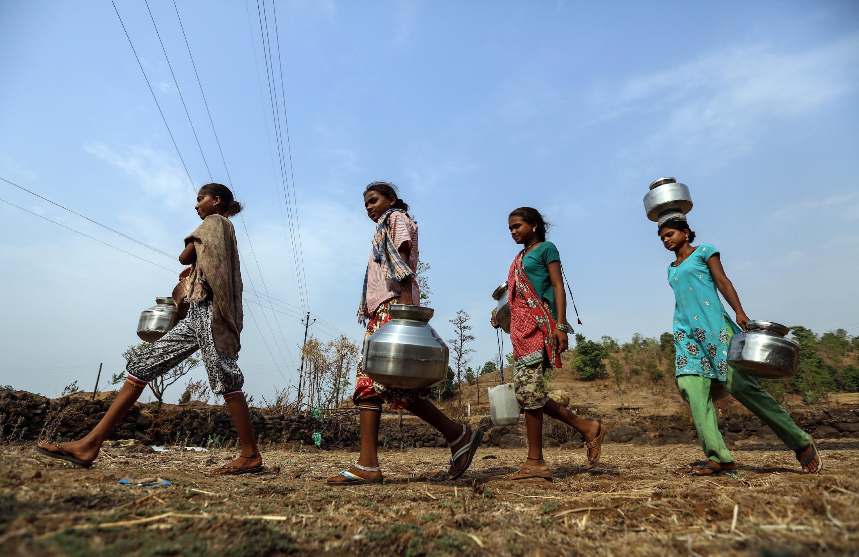 Vier indische junge Frauen gehen in einer Reihe und tragen silbern glänzende Wasserbehälter.