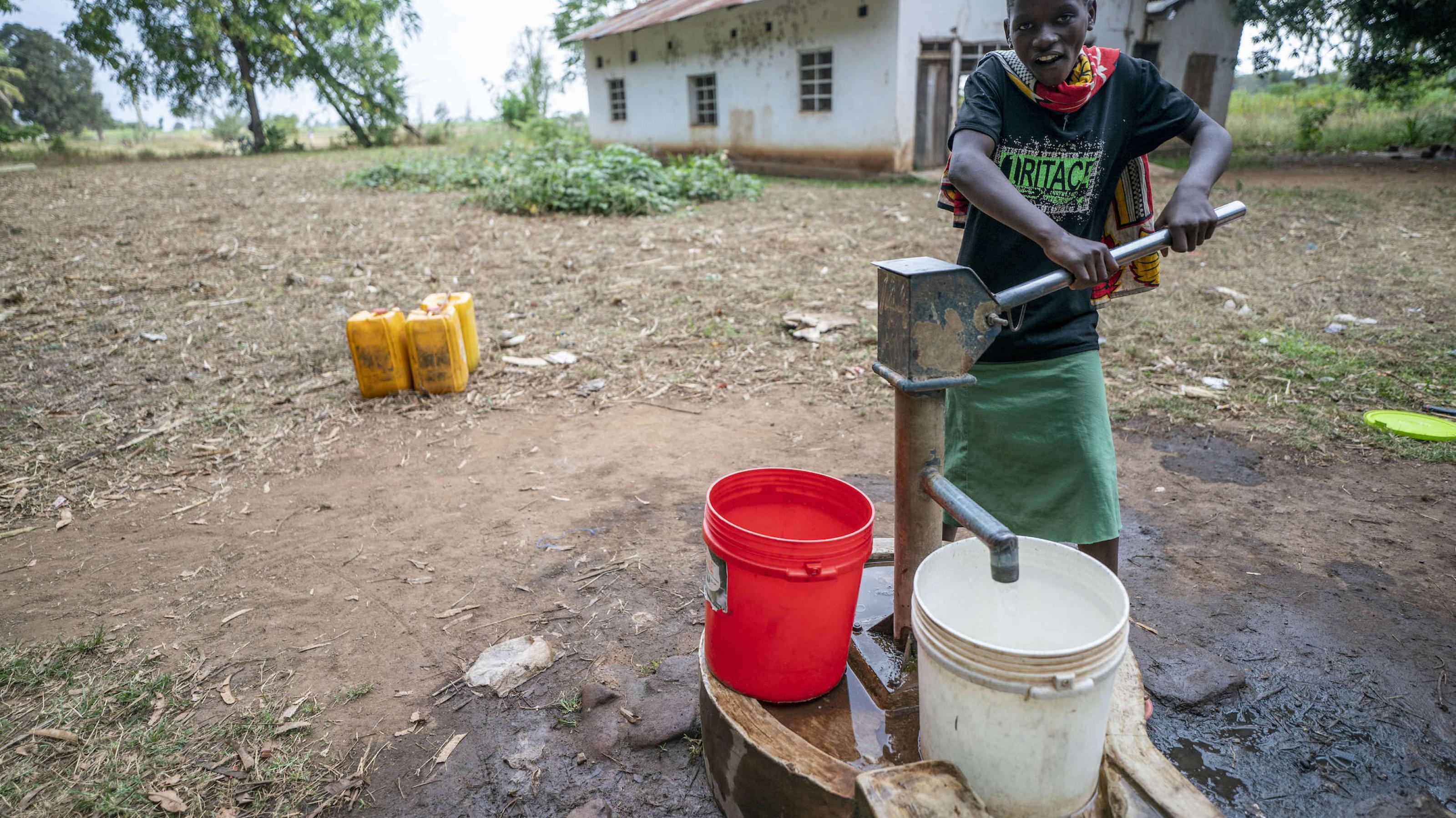 Eine junge Frau holt sich Trinkwasser mit einer sehr einfach gebauten Pumpe. Hinter ihr ein sehr ärmlich wirkendes Haus.
