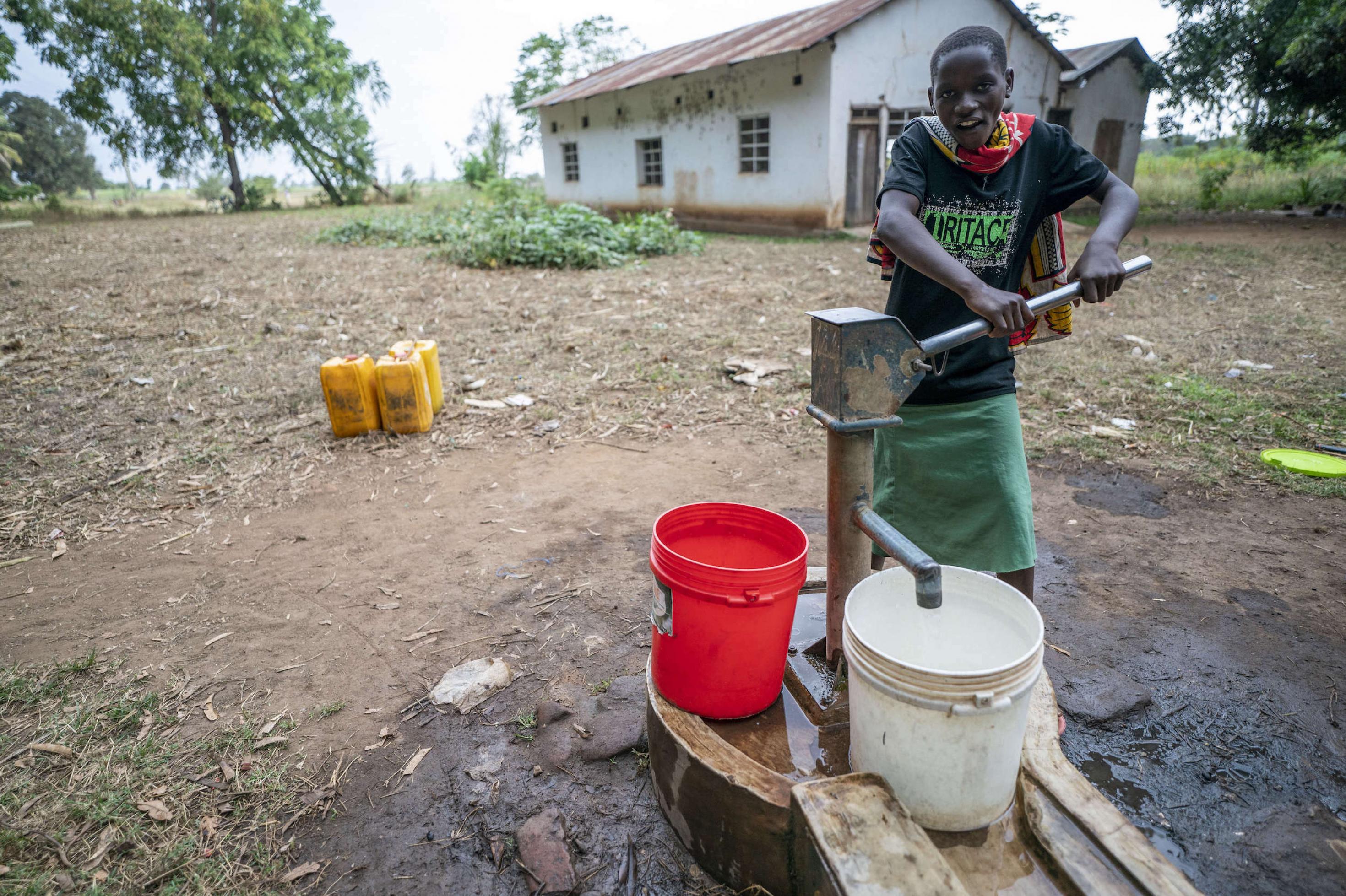 Eine junge Frau holt sich Trinkwasser mit einer sehr einfach gebauten Pumpe. Hinter ihr ein sehr ärmlich wirkendes Haus.