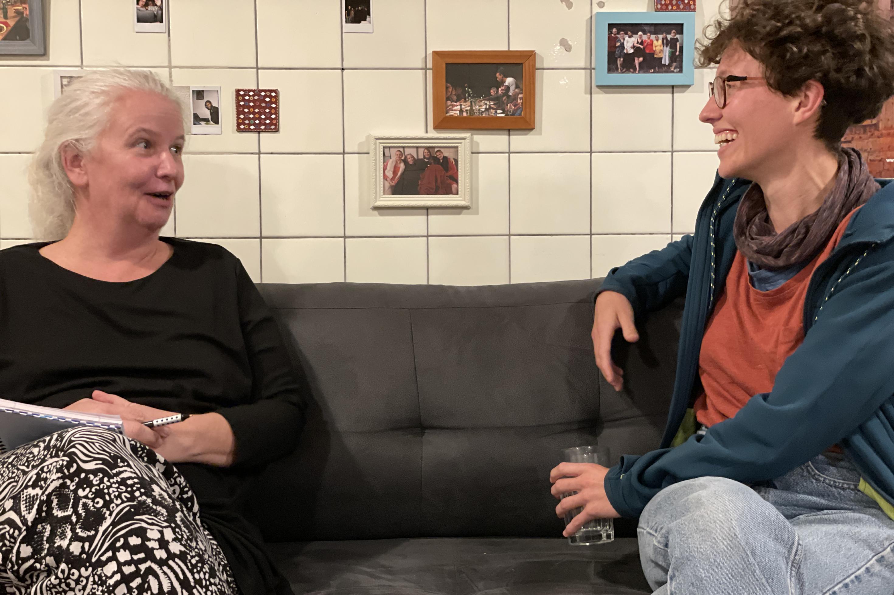 Die beiden Regisseurinnen Barbara Kölling und Clara Gohmert sitzen sich auf einer Couch gegenüber und unterhalten sich. Im Hintergrund weiße Kacheln und viele kleine, gerahmte Bilder.