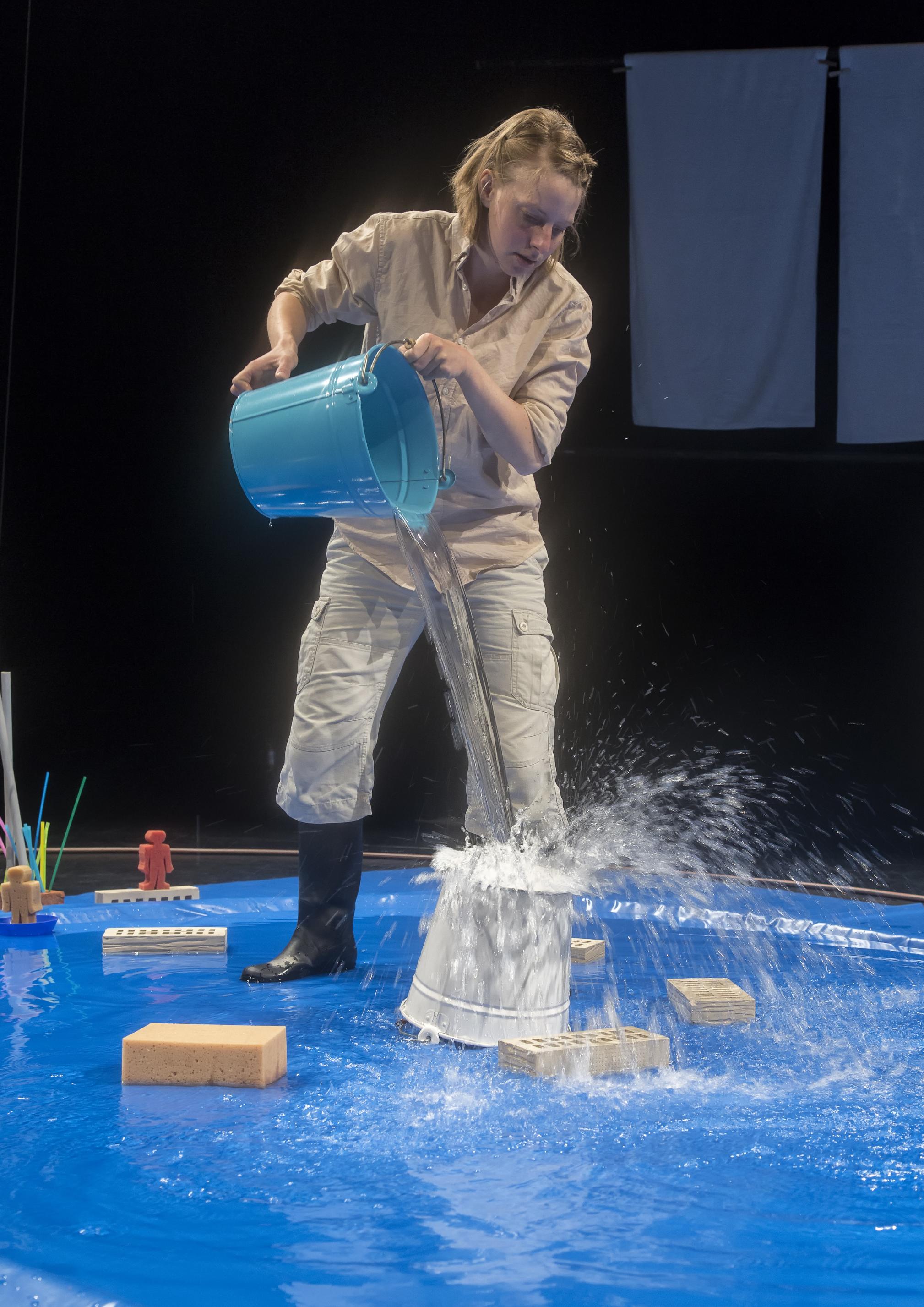 Eine Frau steht in Gummistiefeln in einem kleinen, künstlichen Teich auf der Bühne und lässt Wasser aus einem blauen Eimer auf den Boden eines umgedrehten Eimers fallen. Schwungvoll spritzen die Tropfen wieder hoch und von dort in den Teich.