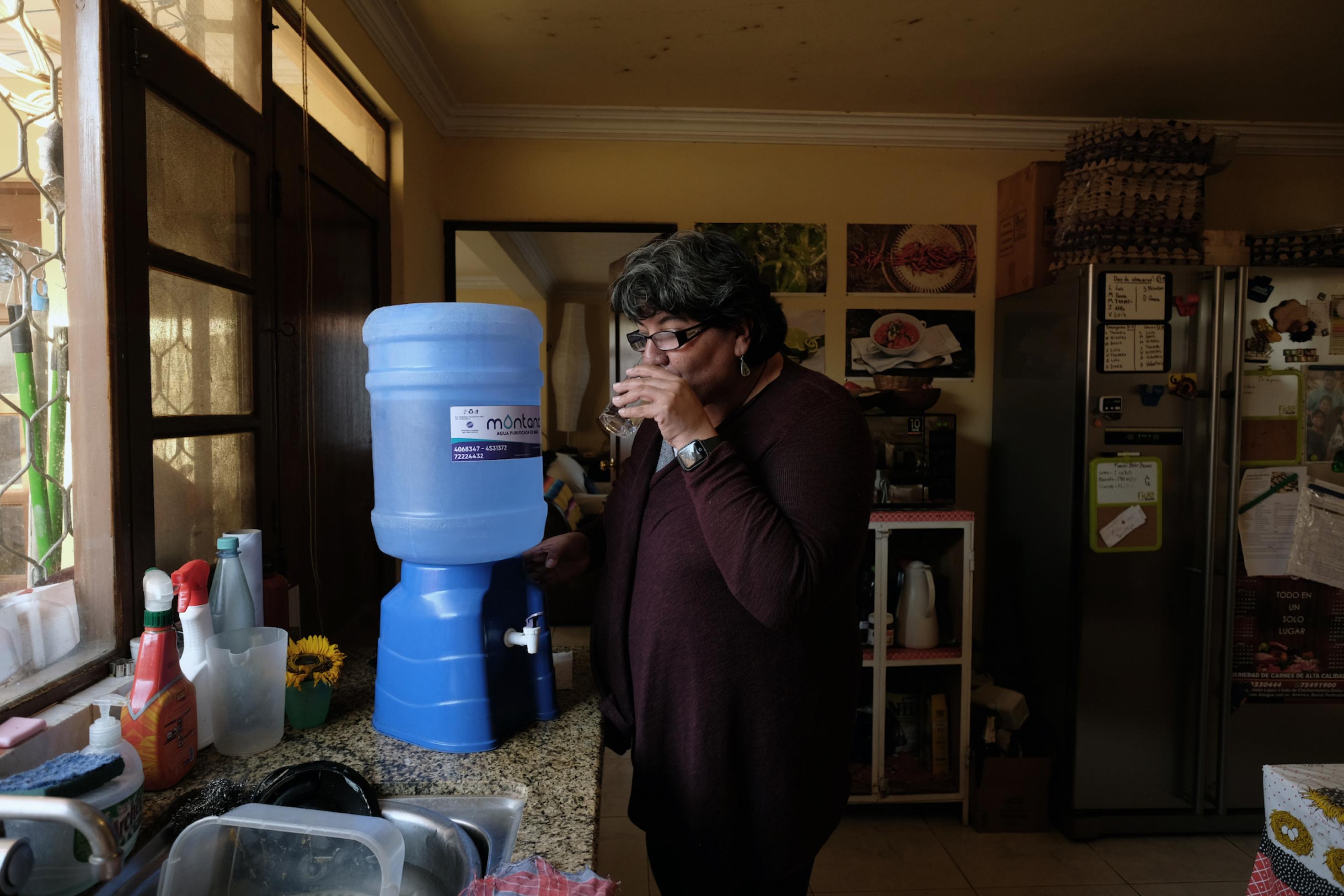 Frau steht in Küche neben Wasserspender und trinkt ein Glas Wasser.