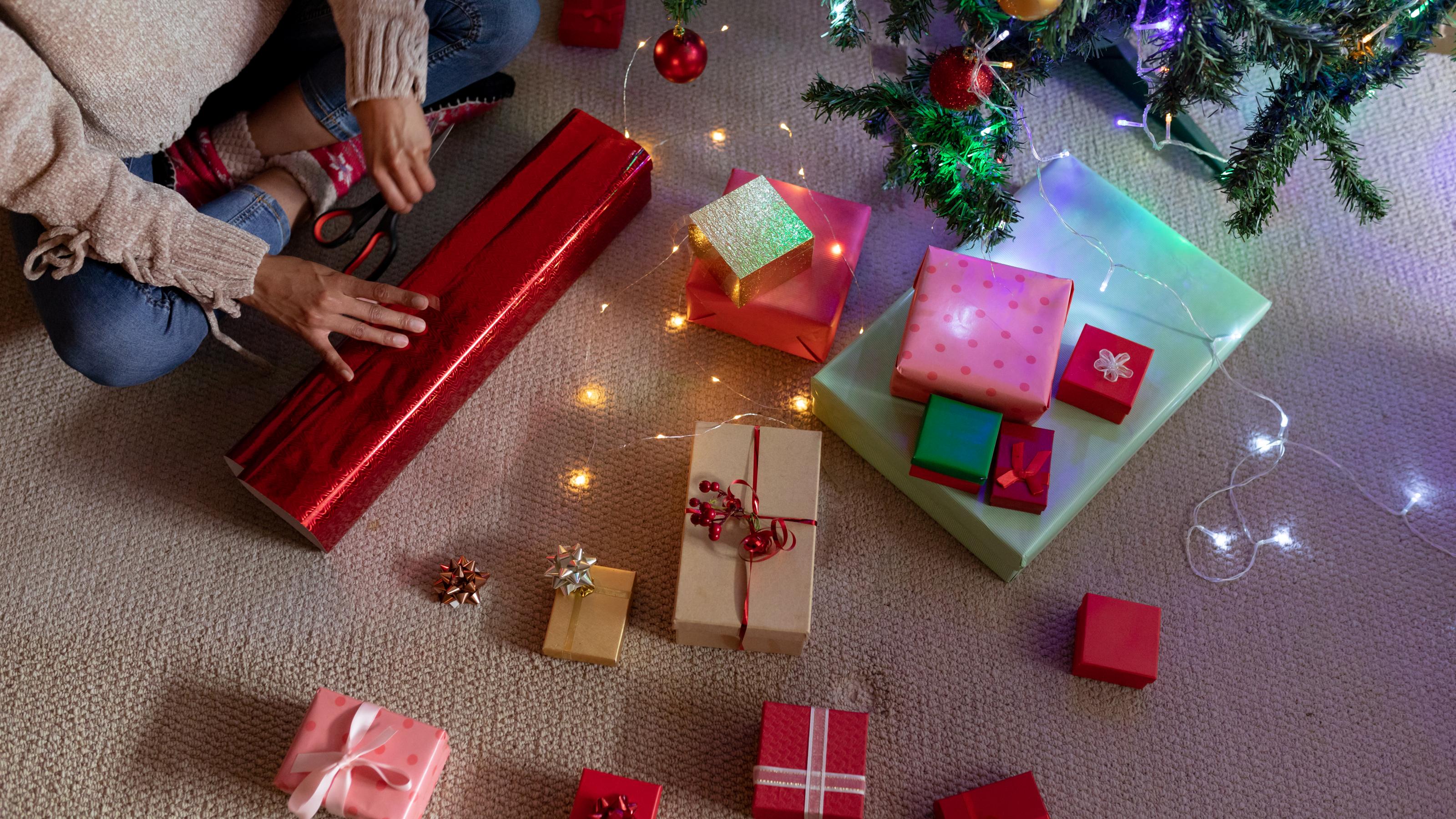 Eine Frau sitzt im Schneidersitz auf dem Boden und verpackt ein Geschenk in rotes Geschenkpapier. Vor ihr auf dem Boden liegen bereits fertig verpackte Geschenke.