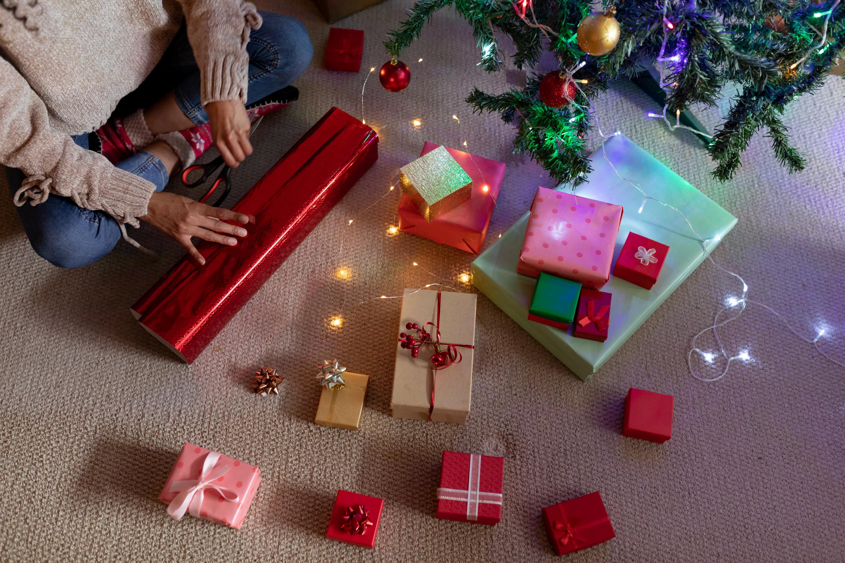 Eine Frau sitzt im Schneidersitz auf dem Boden und verpackt ein Geschenk in rotes Geschenkpapier. Vor ihr auf dem Boden liegen bereits fertig verpackte Geschenke.