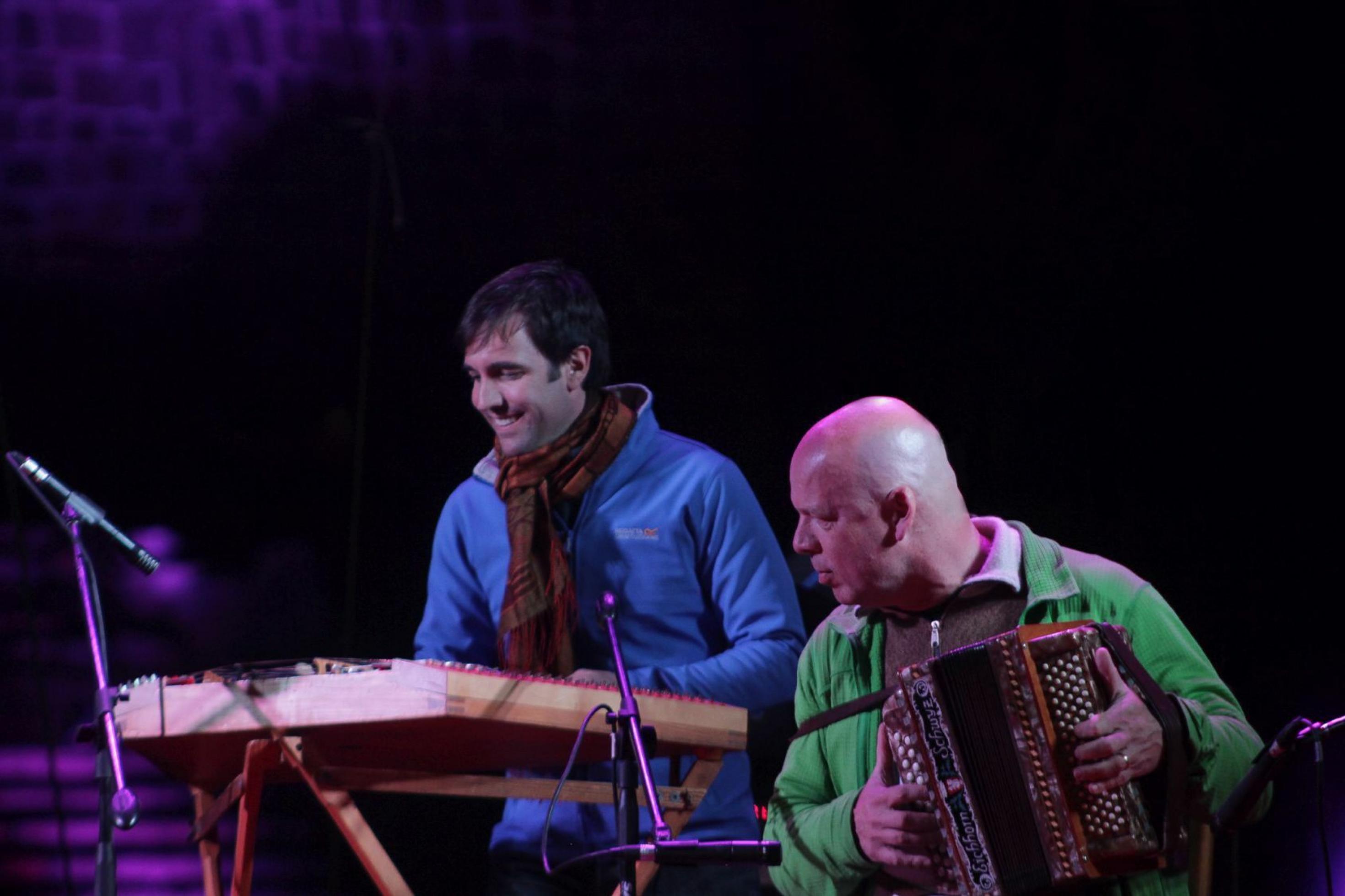 Zwei Männer musizieren auf einer Freilichtbühne, nachts. Links ein Hackbrettspieler, rechts im Vordergrund ein kahlköpfiger Mann mit einem Schweizer Handörgeli