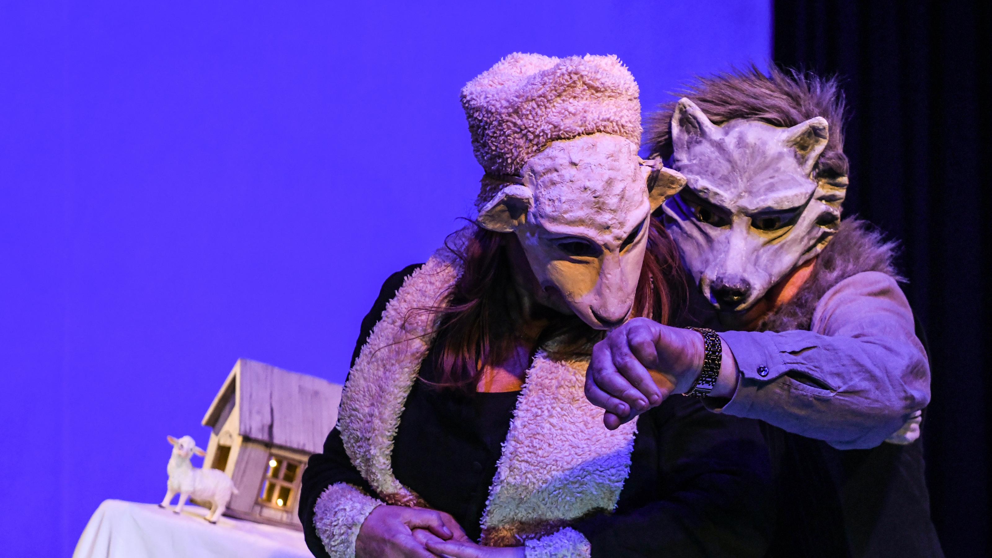 Crischa Ohler und Sjef van der Linden spielen das Schaf und den Wolf. Sie tragen plastische Masken, die die Gesichter halb bedecken. Der Wolf zeigt dem Schaf gerade seine teure Uhr.
