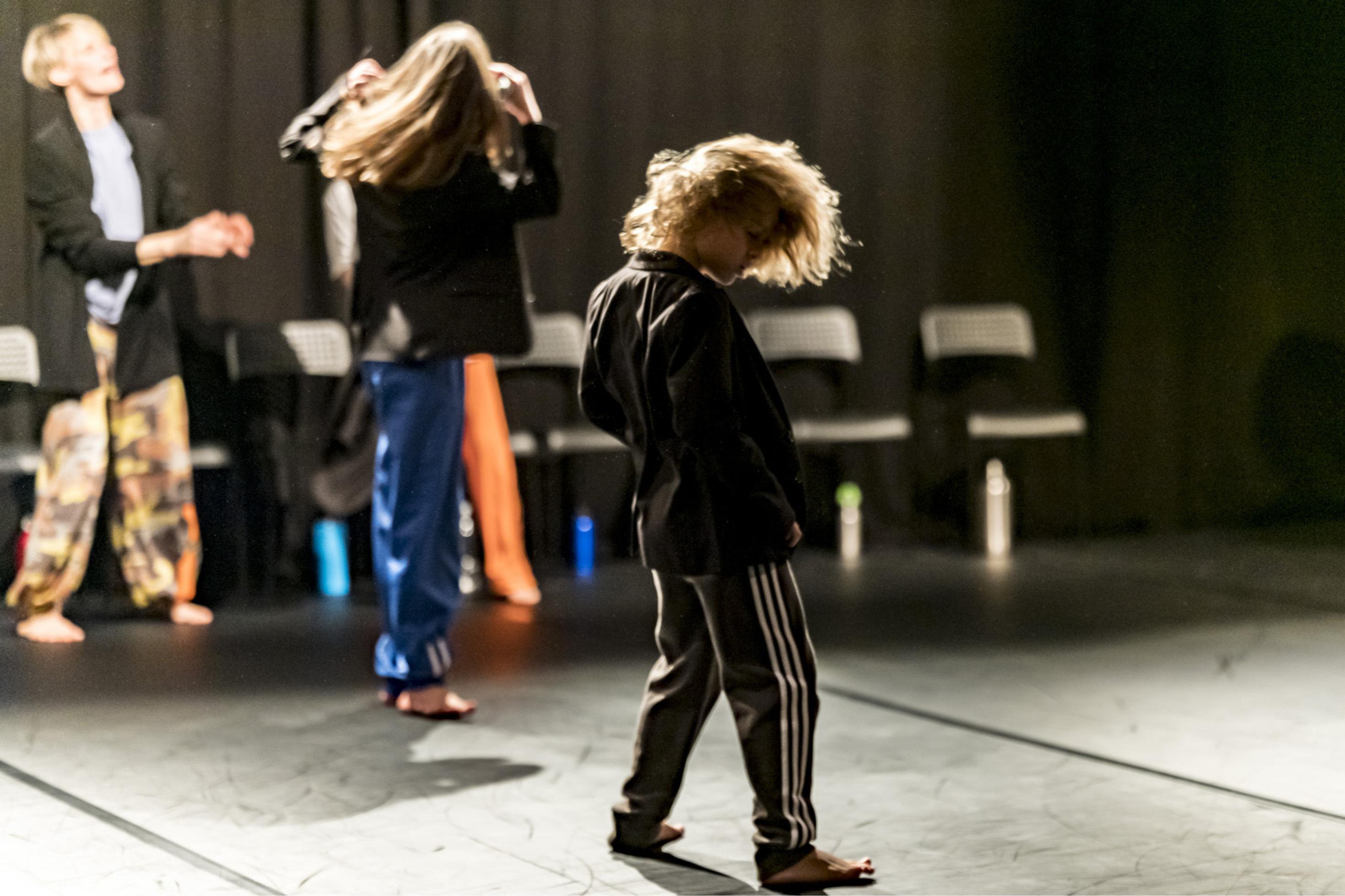 Auf der Bühne sieht man einen kleinen Jungen wild tanzen. Im Hintergrund bewegen sich noch zwei weitere Menschen, ein sehr junger so schnell, dass die Konturen verwischen. Es ist eine Szene aus dem Tanzstück „Mischpoke“ von Barbara Fuchs.