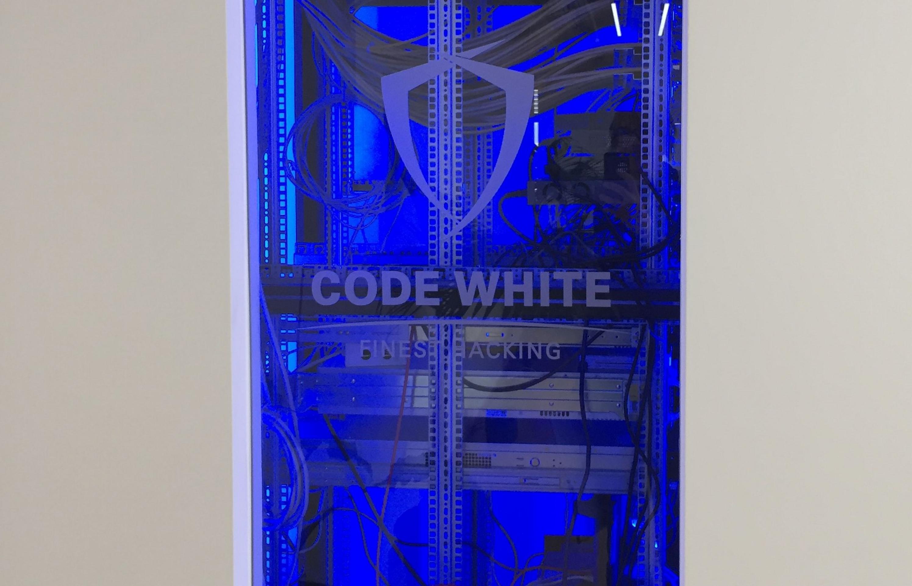 Ein großer Serverschrank, auf dessen Glastür das Firmenlogo eingraviert ist, wurde in eine weiße Wand eingelassen. Das Innere des Schrankes ist komplett in kaltes blaues Licht getaucht. In ihm befindet sich, umgeben von einer Menge Kabel, ein wuchtiger Hochleistungscomputer in einem unspektakulären Metallgehäuse.