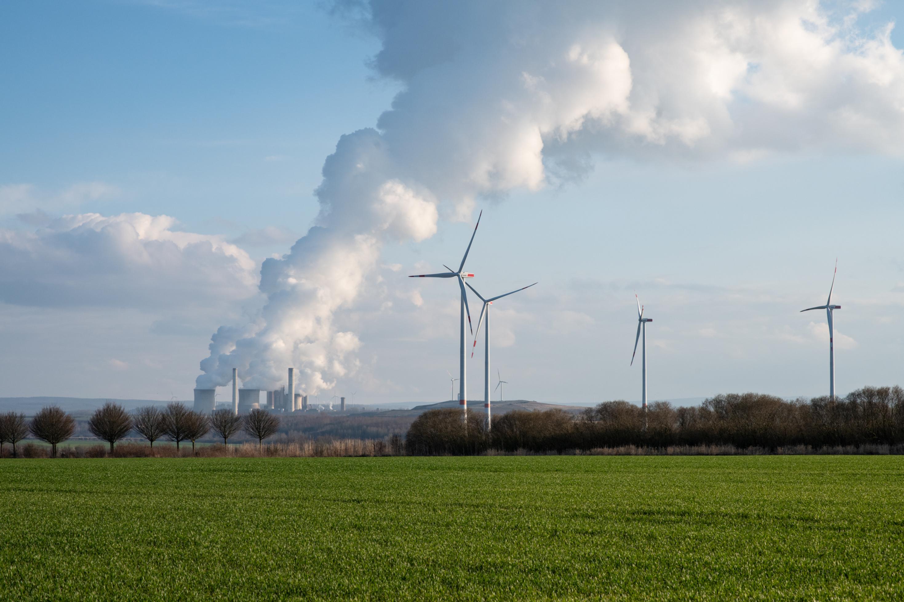 Im Hintergrund qualmt ein Kohlekraftwerk, im Vordergrund stehen mehrere Windkraftanlagen.