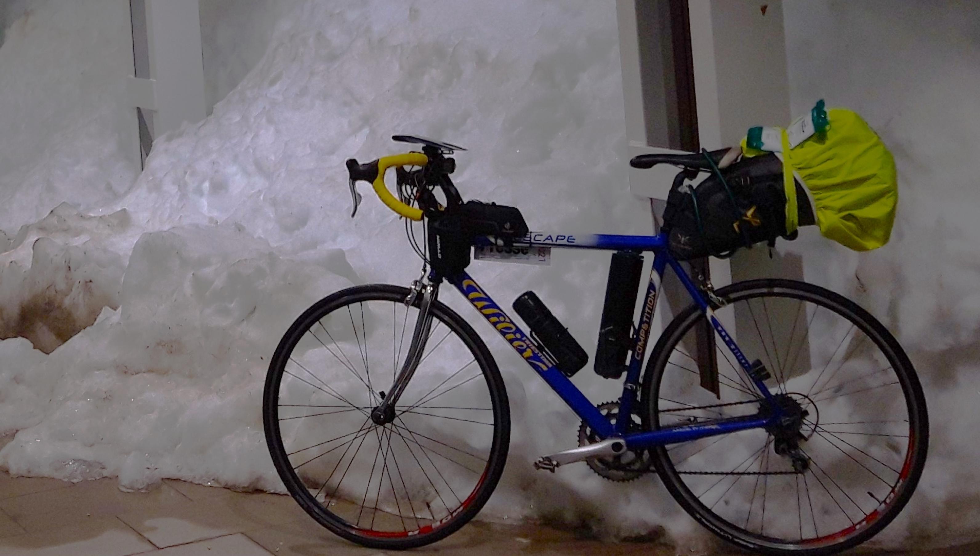 Das Reise-Rennrad des RadelndenReporters lehnt im Dunkeln an einer Bahnhofssäule, hinter der ein etwa zwei Meter hoher Schneewall aufragt.