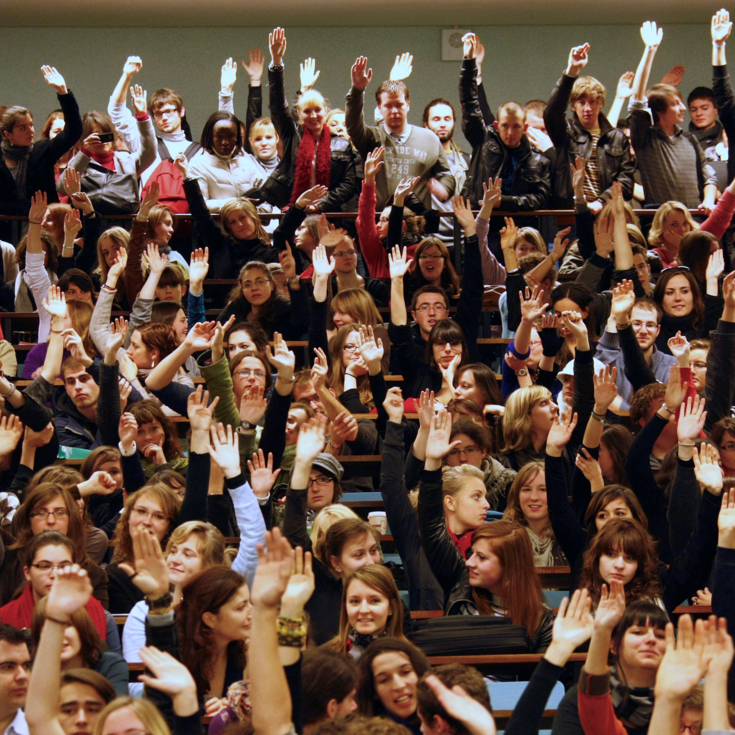 Eine große Zahl von Studierenden im Hörsaal. Sie sitzen dicht gedrängt in den Reihen und heben die Hände bei einer Abstimmung. Die Studierenden sind mehrheitlich weiß.