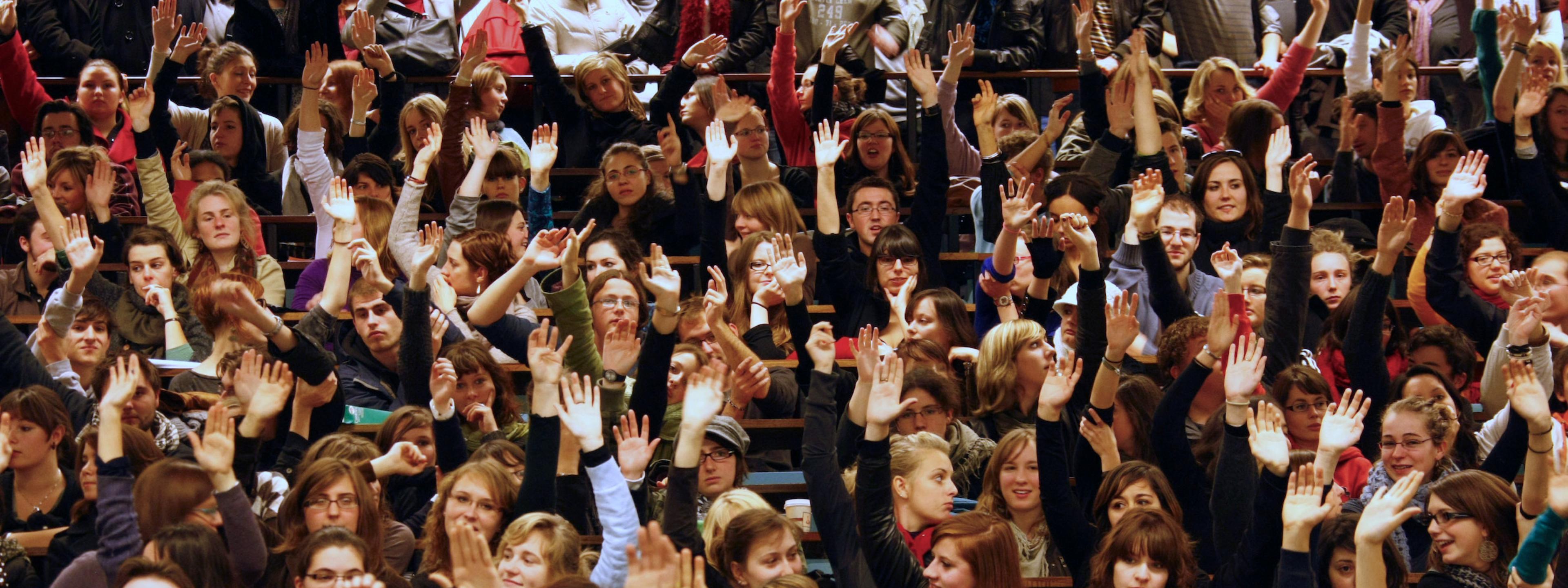Eine große Zahl von Studierenden im Hörsaal. Sie sitzen dicht gedrängt in den Reihen und heben die Hände bei einer Abstimmung. Die Studierenden sind mehrheitlich weiß.