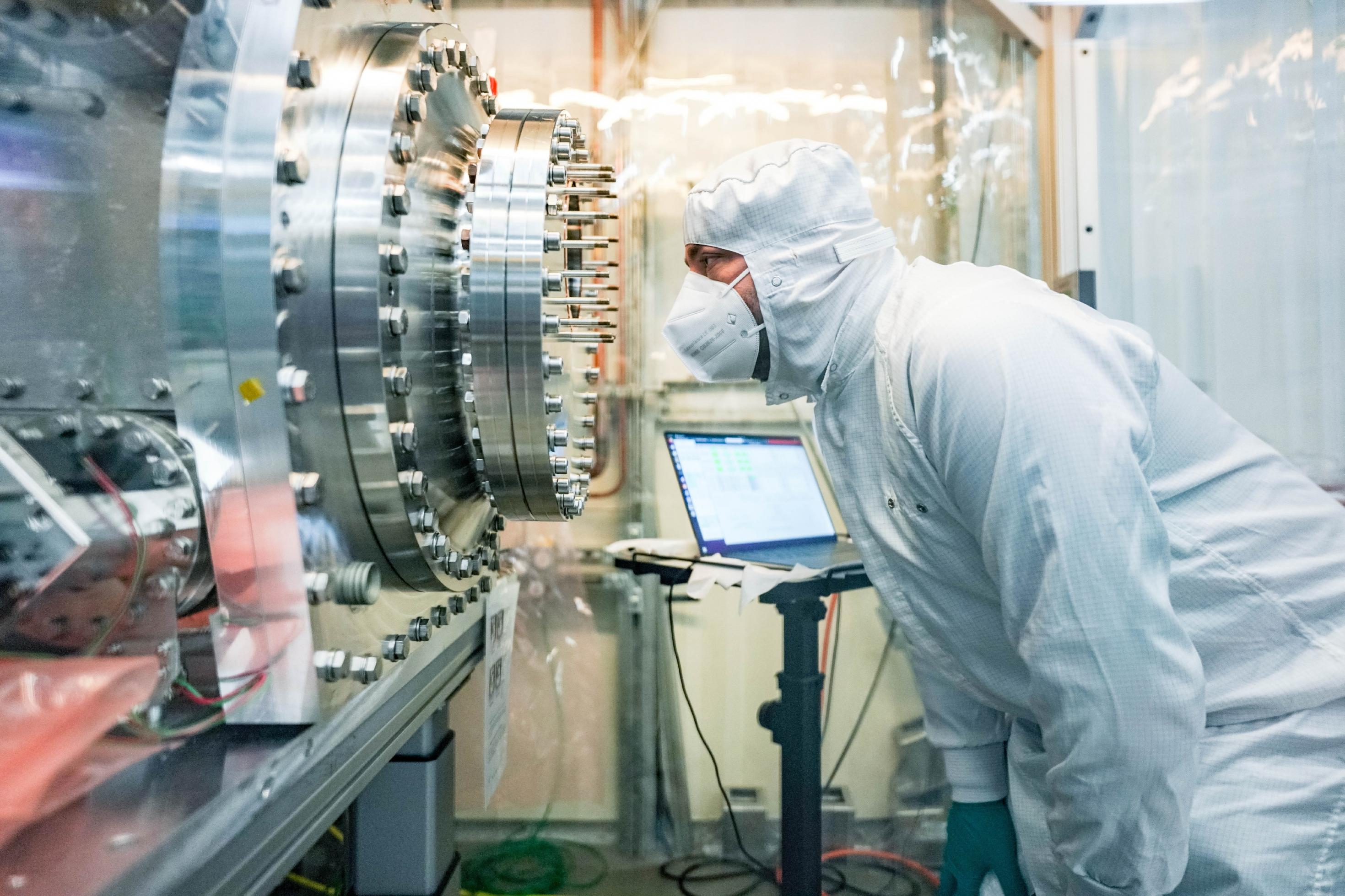 EIn Wissenschaftler in weißem Schutzanzug schaut auf ein kompliziertes metallisches Gebilde.