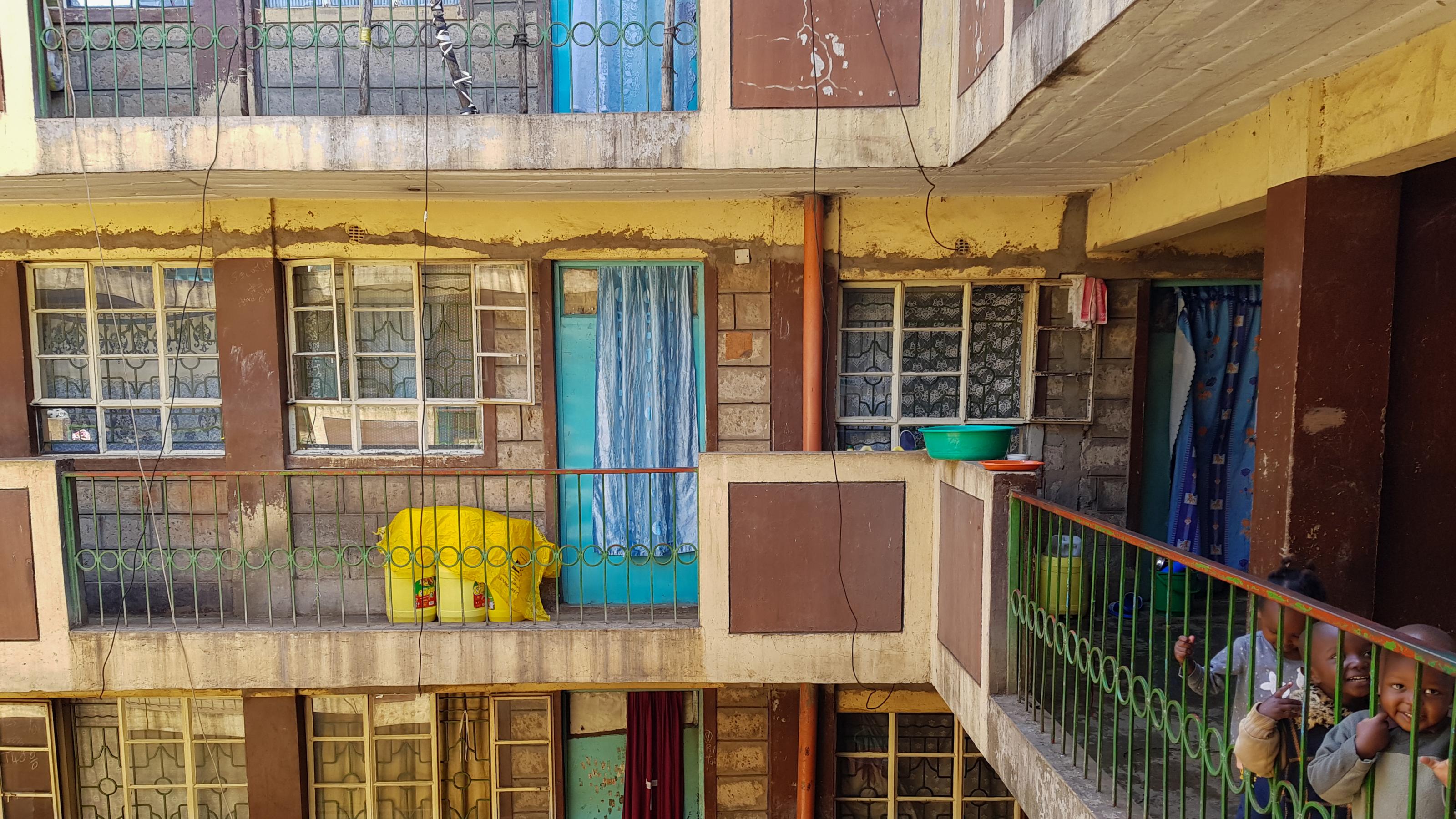 Aufnahme aus dem Innenhof eines Wohnblocks. Rechts am Geländer stehen drei Kinder, die in die Kamera lächeln.
