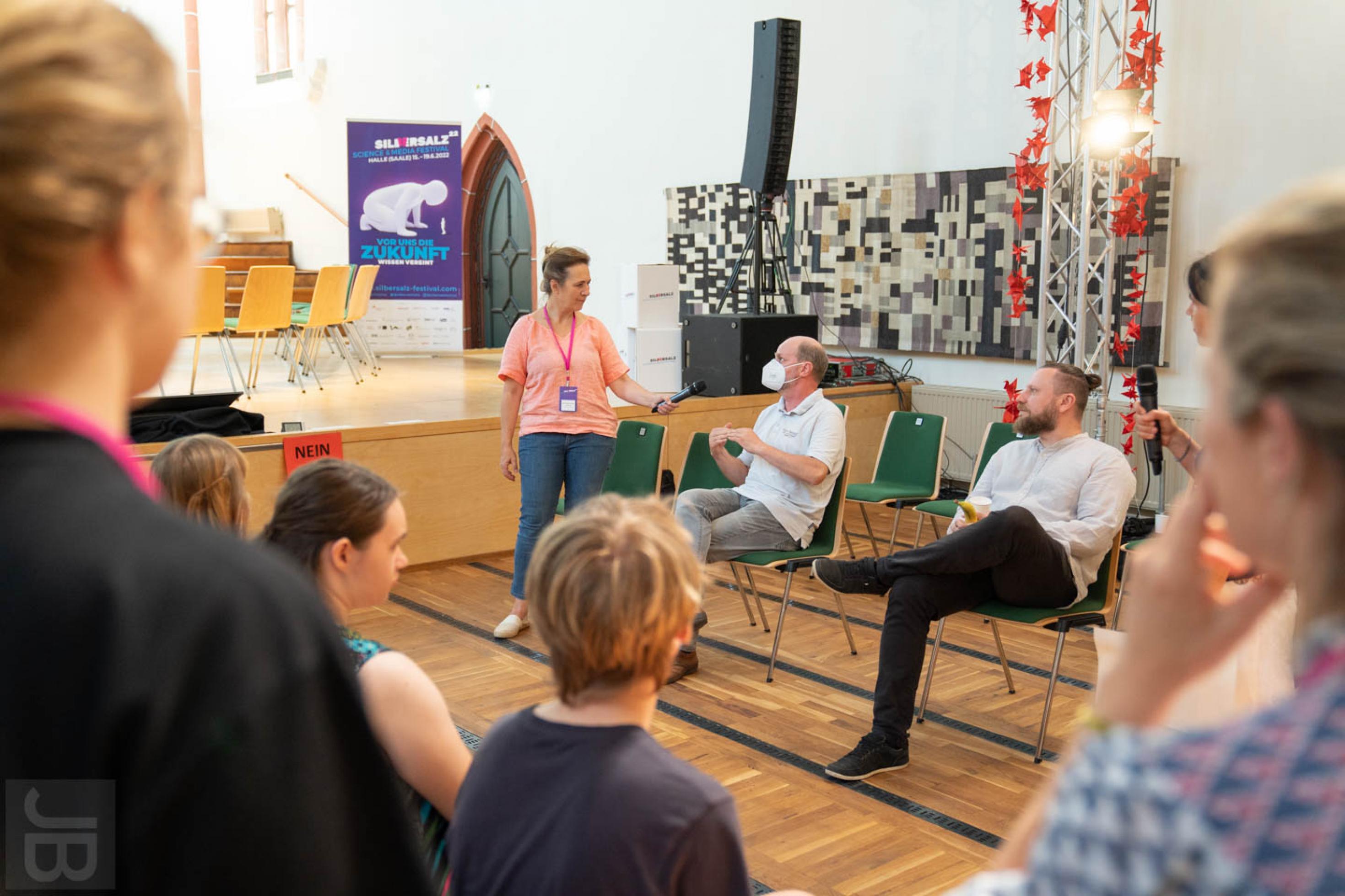 Die Moderatorin hält einem Workshop-Teilnehmer das Mikrofon hin. Er begründet seine Sitzplatzwahl und führt die Argumente aus.