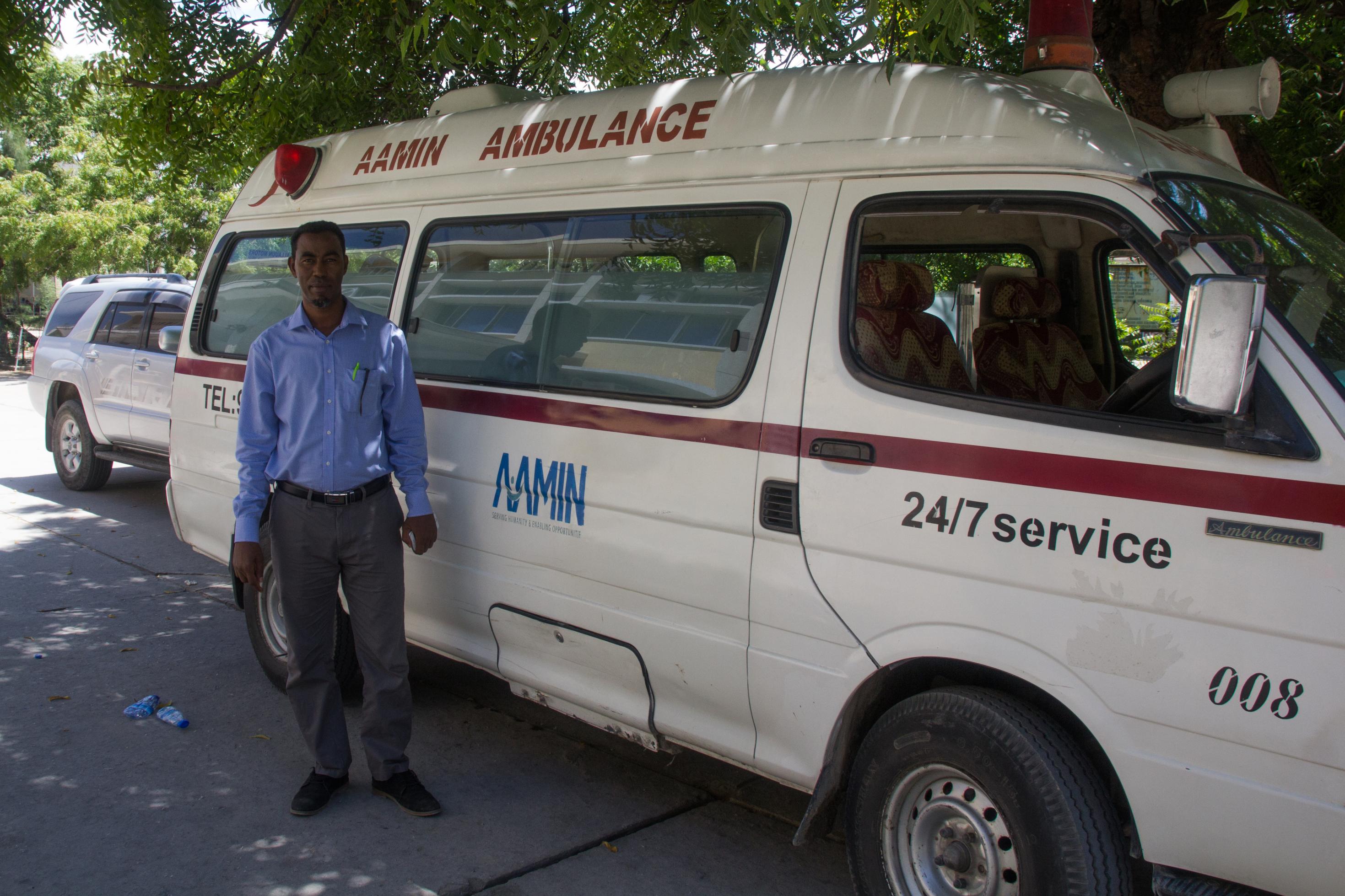 Ein Mann mittleren Alters steht vor einem Rettungswagen. Dieser trägt die Aufschriften „Aamin Ambulance“ und „24/7 Service“.