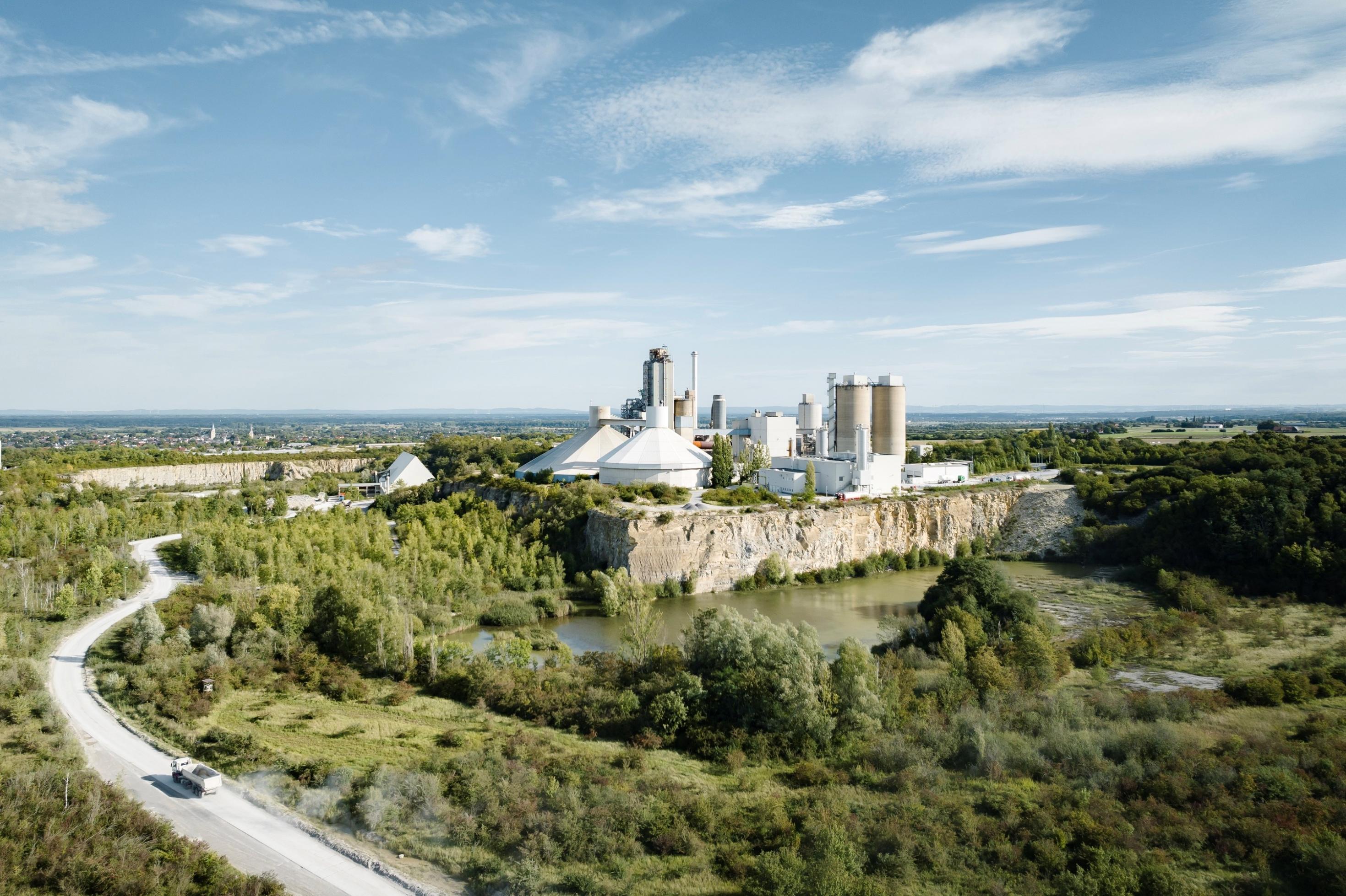 Das Bild zeigt eine Industrieanlage an der Kante eines Steinbuchs. Davor liegt ein mit Bäumen umgebener See. Links im Vordergrund fährt ein LKW auf das Werk zu.