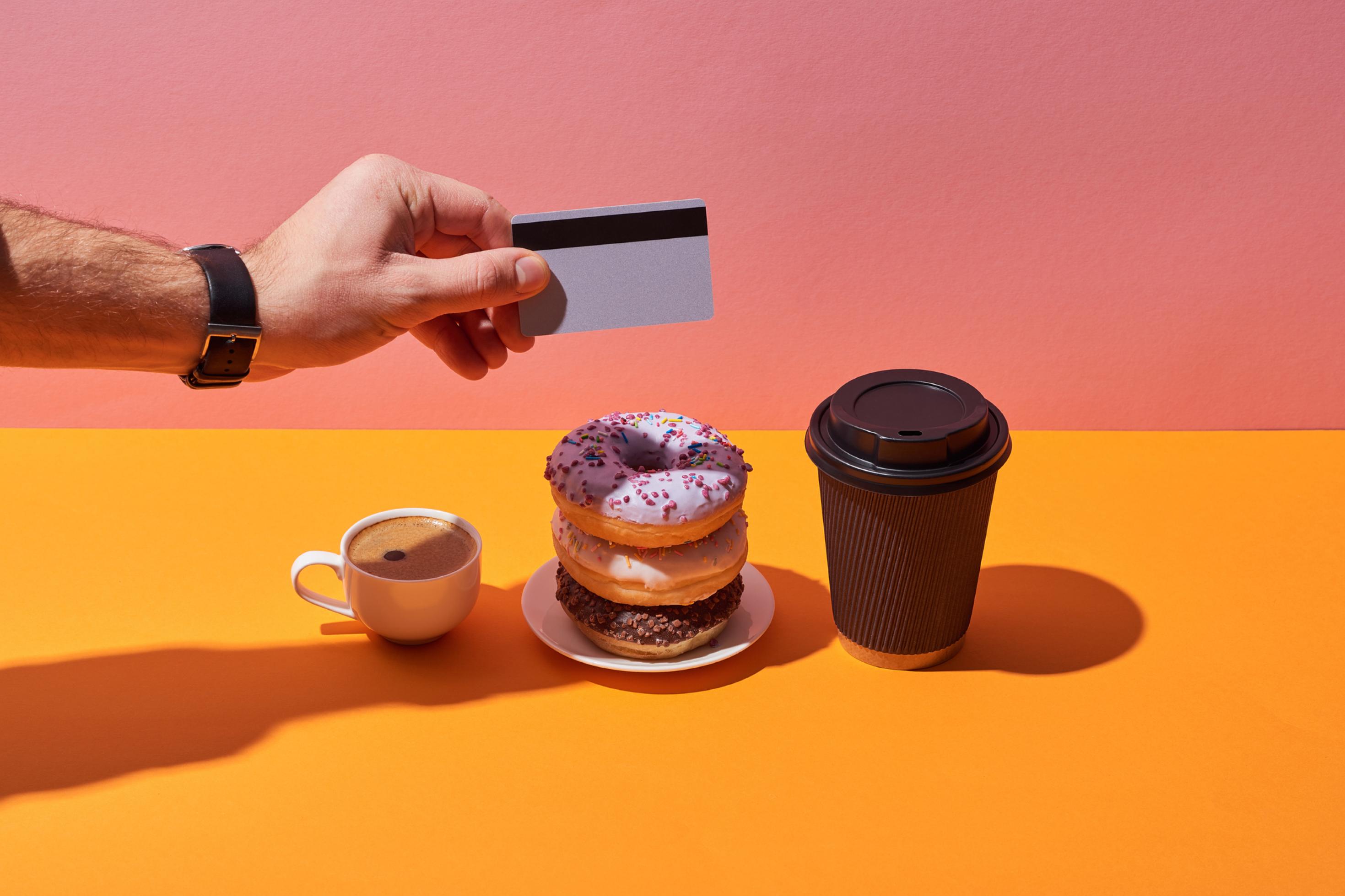 Ein Espresso, drei übereinander gestapelte bunt verzierte Donuts und ein Coffe-to-go stehen nebeneinander. Eine Männerhand hält eine Kreditkarte zum Bezahlen hin.