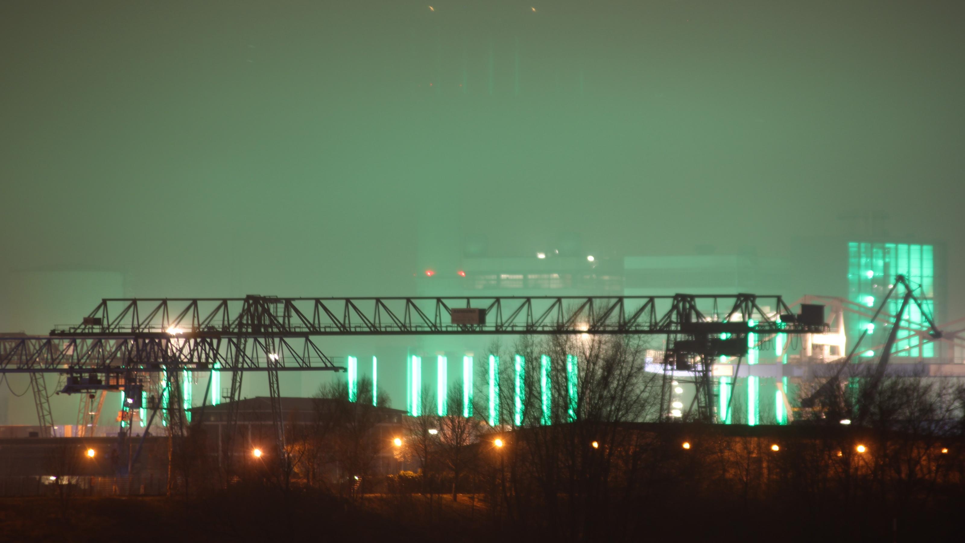 Die grüne Beleuchtung eines Kraftwerks leuchtet eine abendliche Szene am Düsseldorfer Hafen aus.