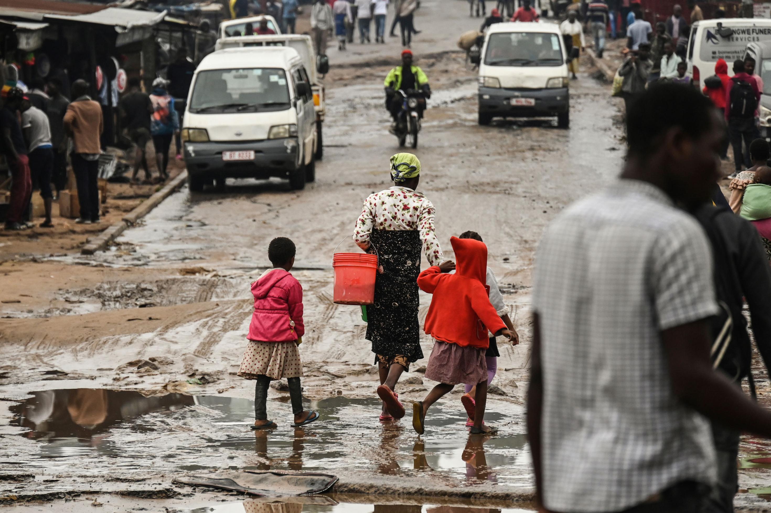 Die Strasse ist nass, teils sind Pfützen zu sehen, eine Frau und zwei Kinder gehen dort entlang, außerdem andere Passanten und Minibustaxis.